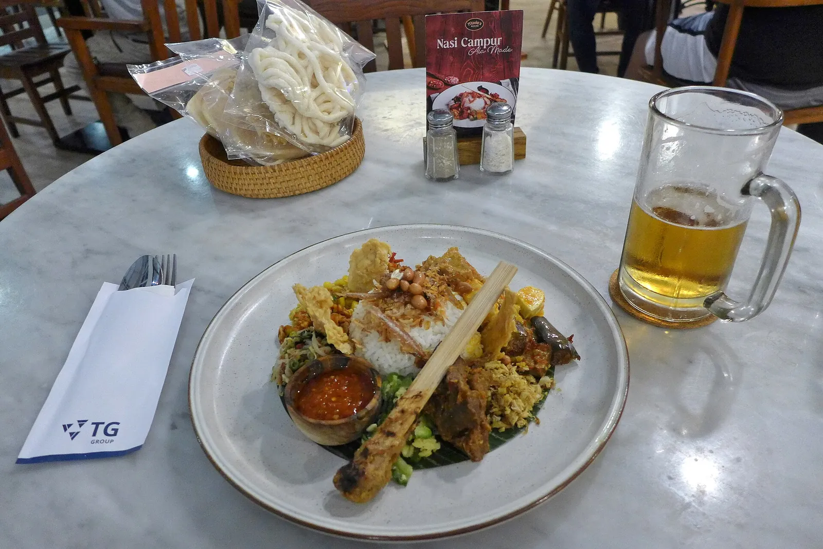 Plate of nasi campur with rice, sate lilit, sambal and crackers next to a glass of beer on a restaurant table