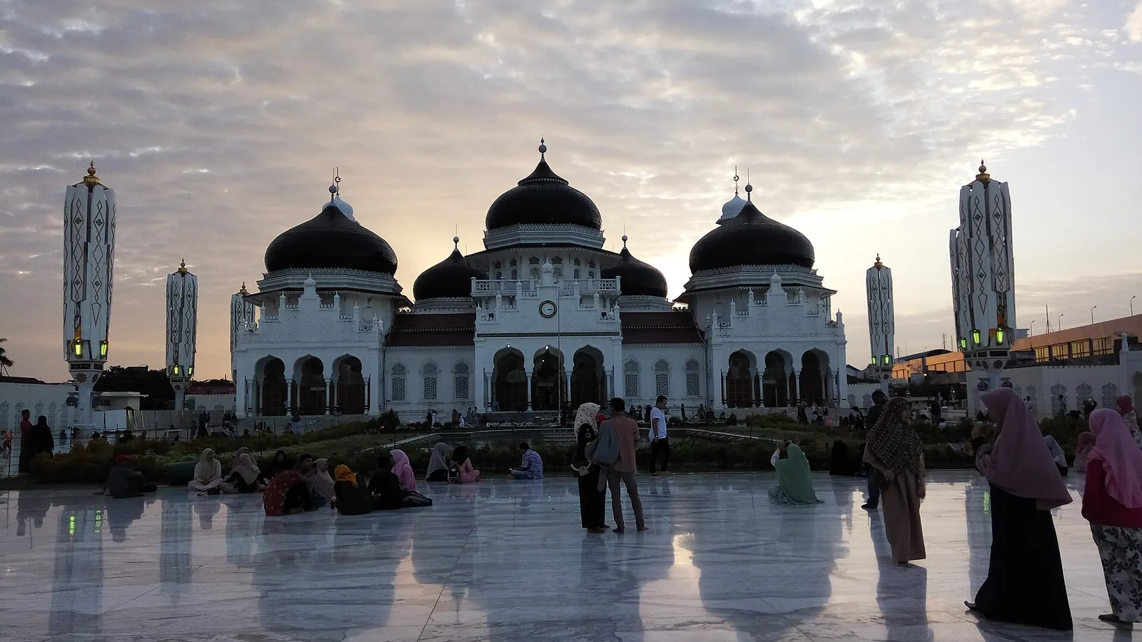 The white Baiturrahman Grand Mosque in Banda Aceh at dusk, with women in headscarves and long robes on the marble courtyard