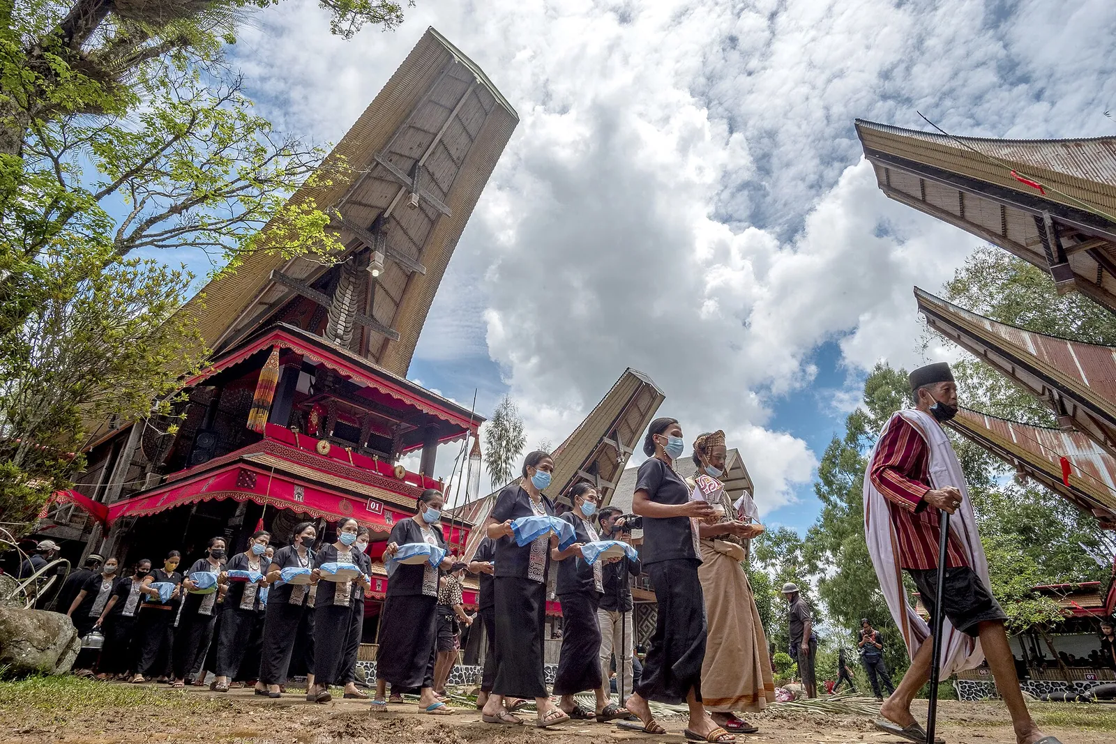 Women dressed in black carrying trays in a line past tongkonan houses, welcoming guests at a Rambu Solo' funeral in Toraja