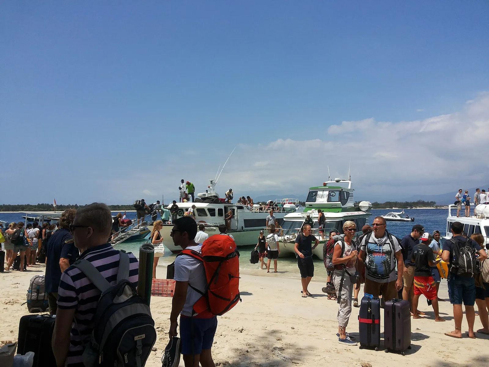 Travellers with backpacks and suitcases on the beach at Gili Trawangan beside moored fast boats