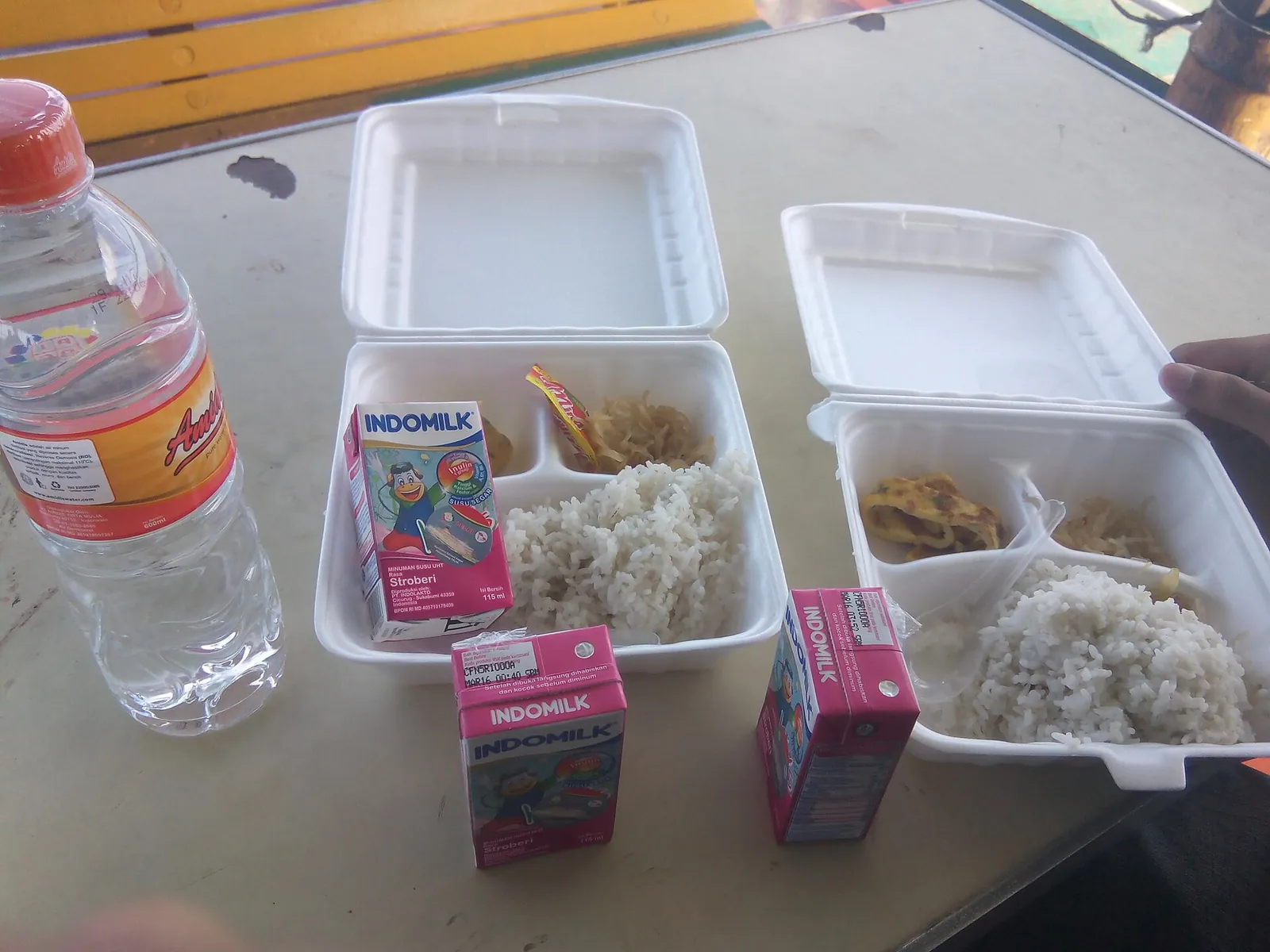 Two foam boxes of rice with side dishes, a bottle of water and cartons of milk on a table aboard a Pelni ship