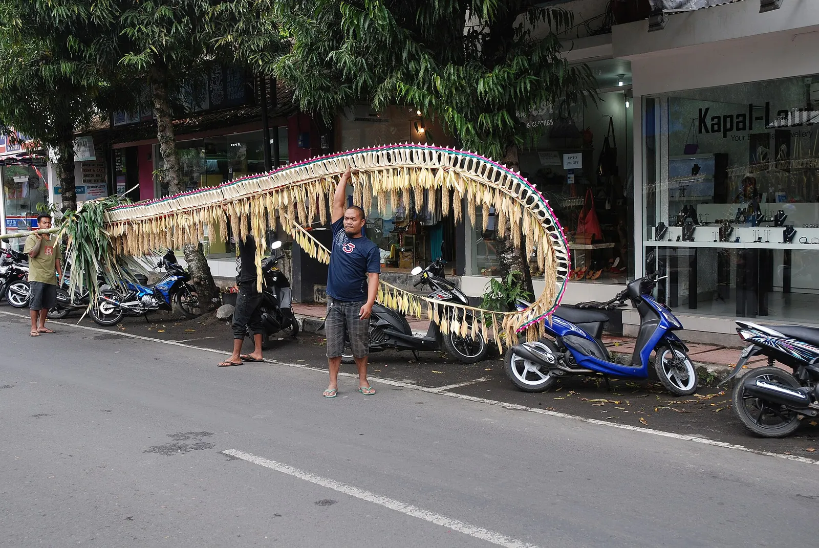 A long curved penjor bamboo pole with woven coconut-leaf decorations by a shop-lined street in Ubud
