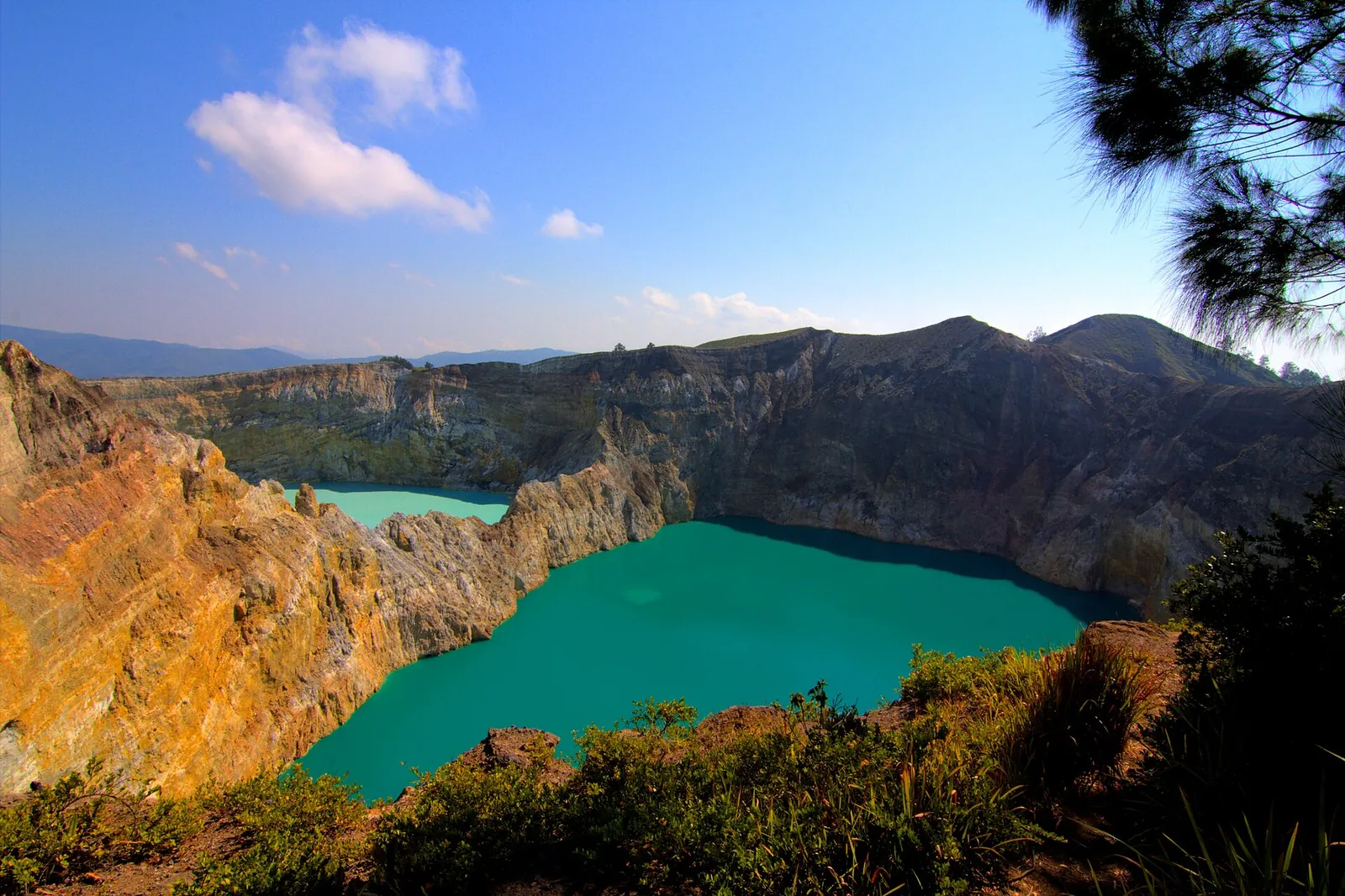 A turquoise crater lake at Kelimutu below eroded orange and grey crater walls under a blue sky