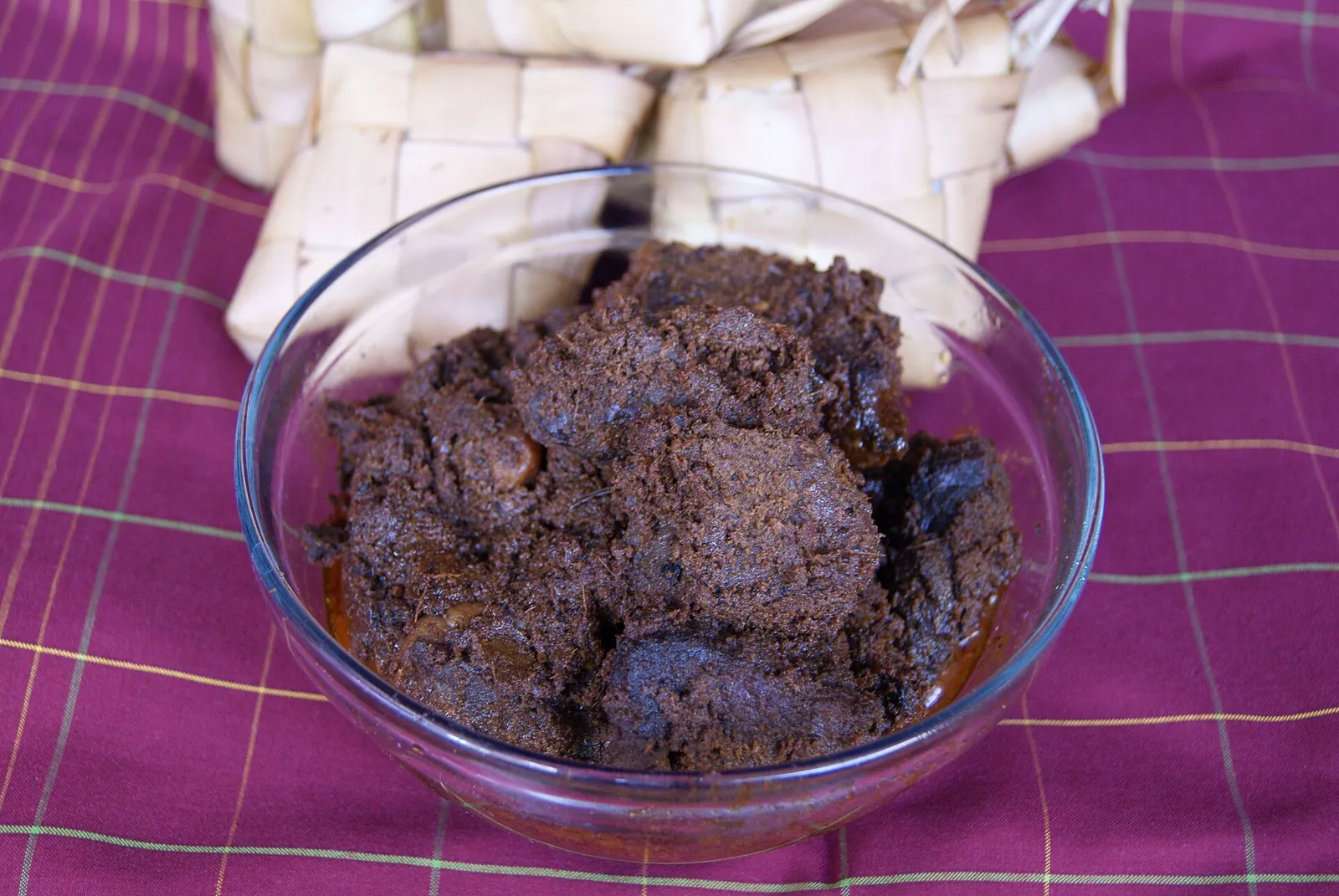 A glass bowl of dark, dry beef rendang with ketupat rice cakes behind it