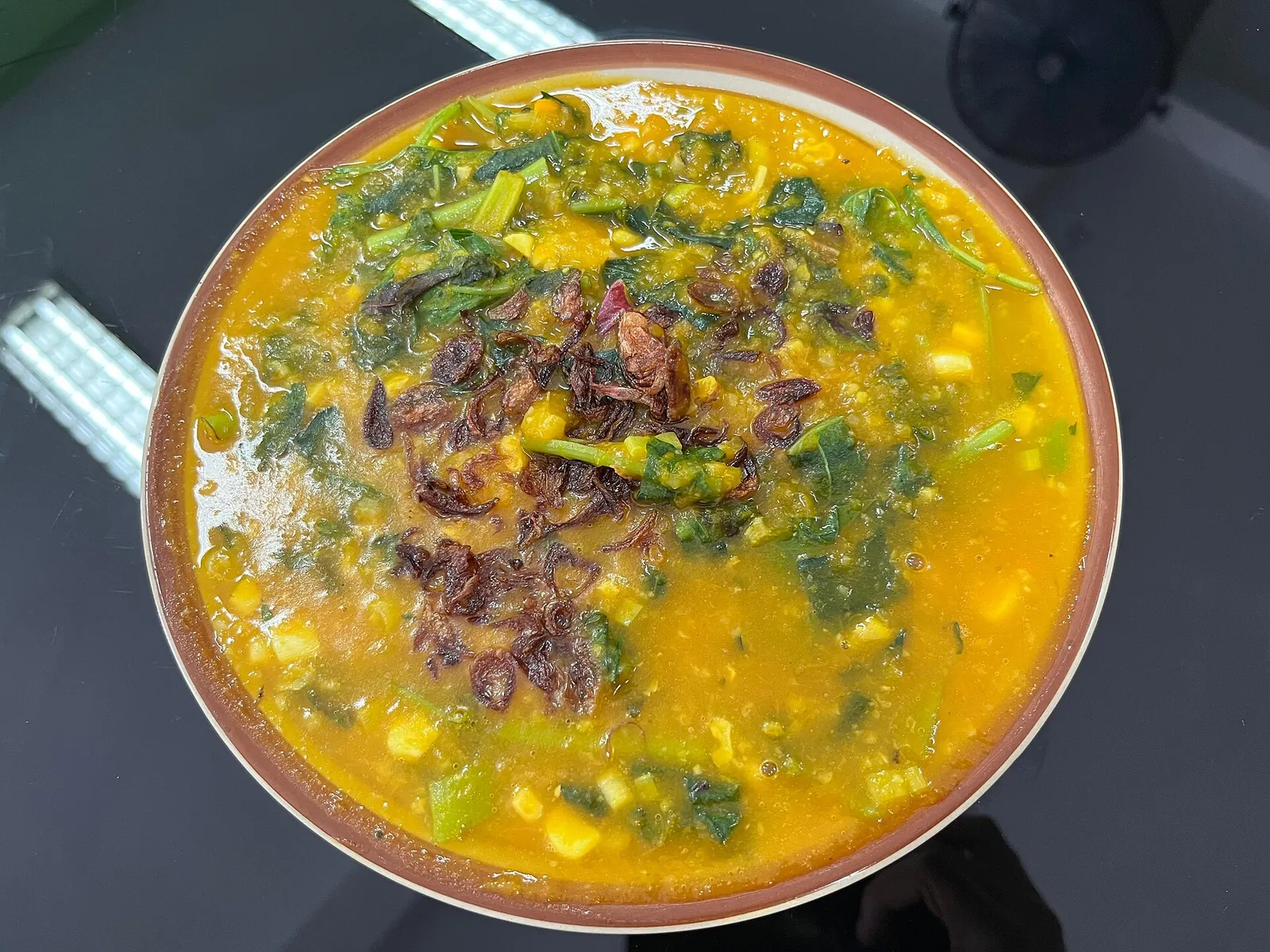 A wide bowl of yellow rice porridge with corn, greens and fried shallots on top