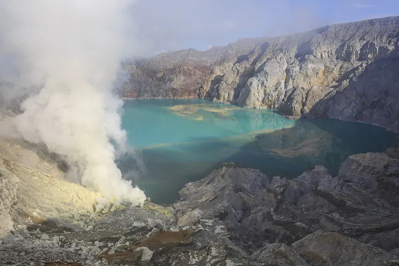 Turquoise acid lake on the floor of Kawah Ijen with white sulphur steam rising from the crater wall
