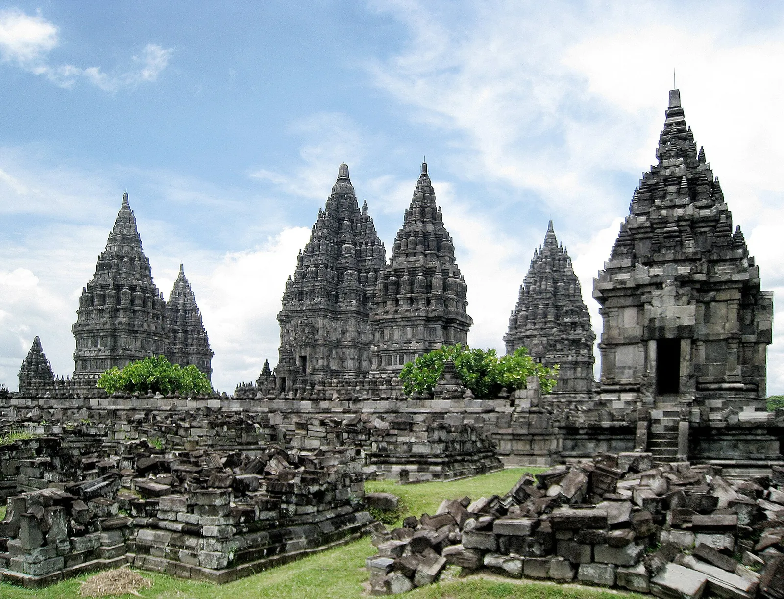 The tall Hindu shrines of Prambanan under a bright blue sky, with fallen stone blocks of smaller shrines in front