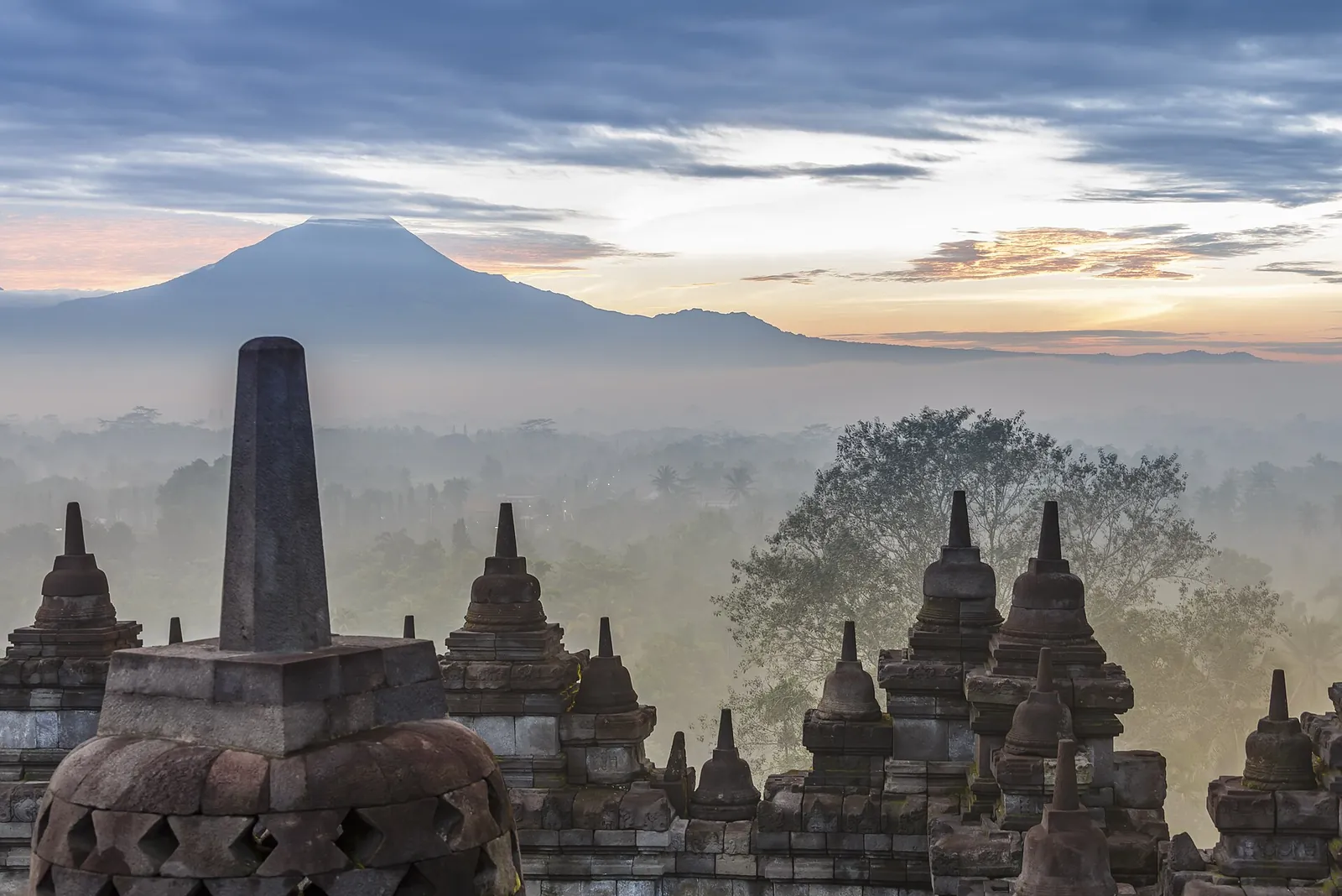 Perforated stupas on Borobudur's upper terrace at dawn, with mist over the plain and a volcano on the horizon