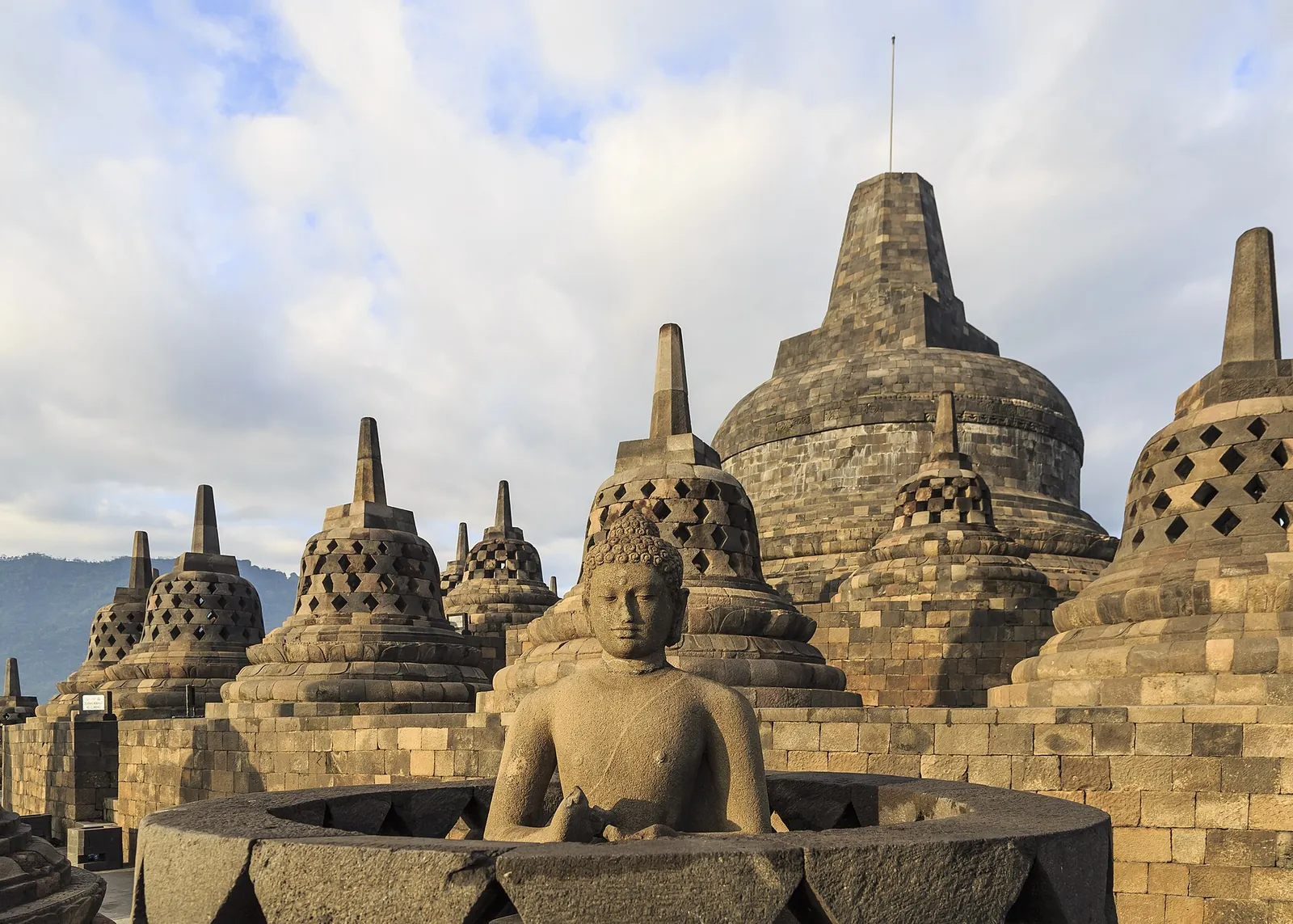 Stone stupas and a seated Buddha on the upper terrace of Borobudur under a blue sky