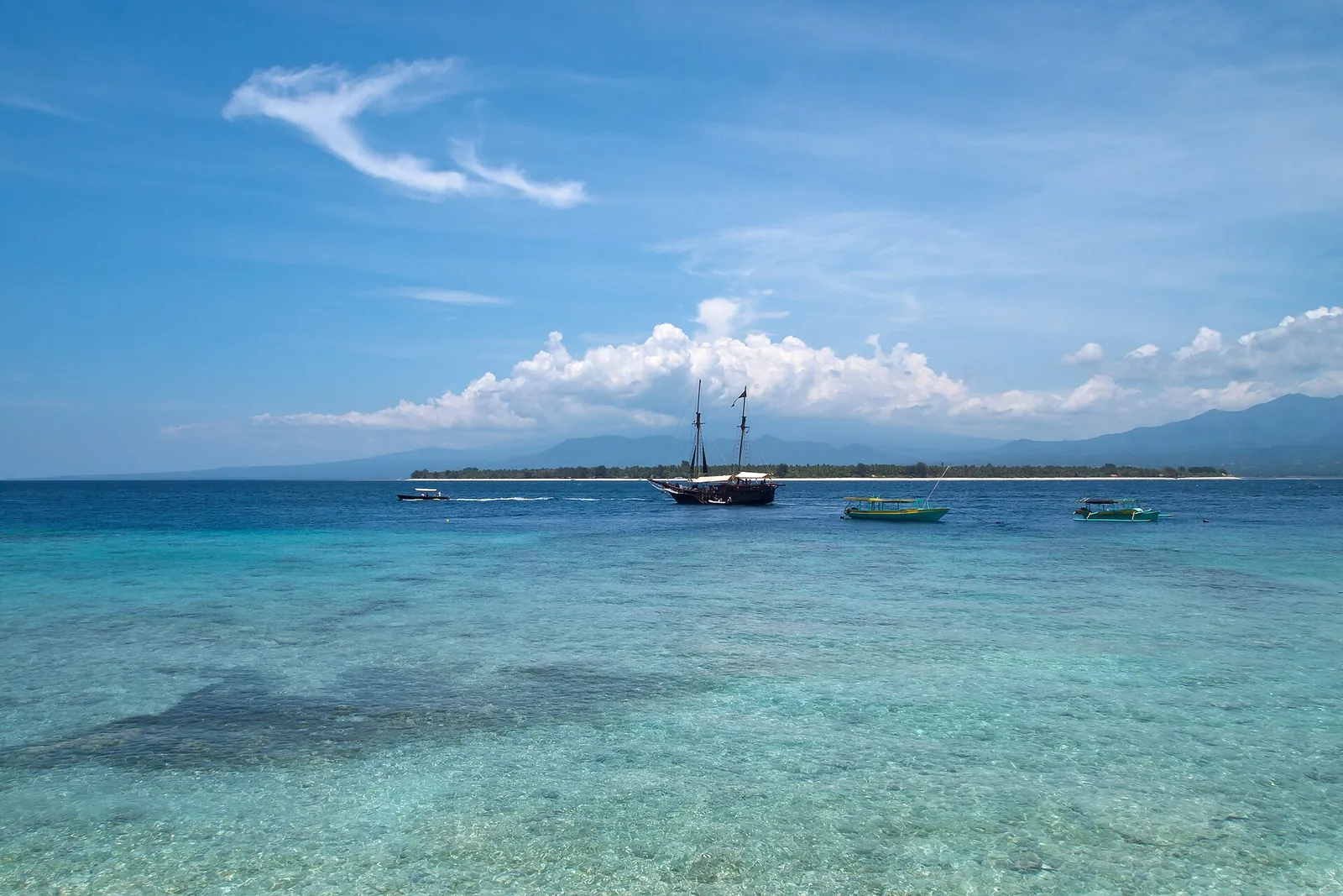 Clear turquoise shallows with Gili Air's low wooded coast and moored boats on the horizon