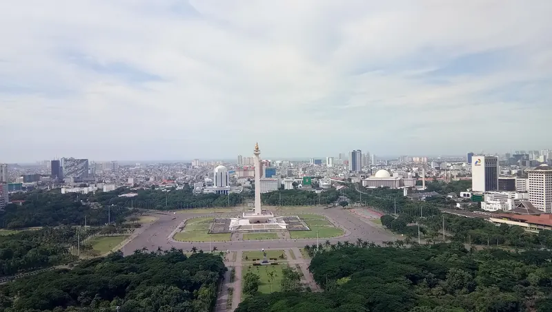 The National Monument (Monas) rising from the green lawns of Merdeka Square in central Jakarta, seen from above with the city spread behind it