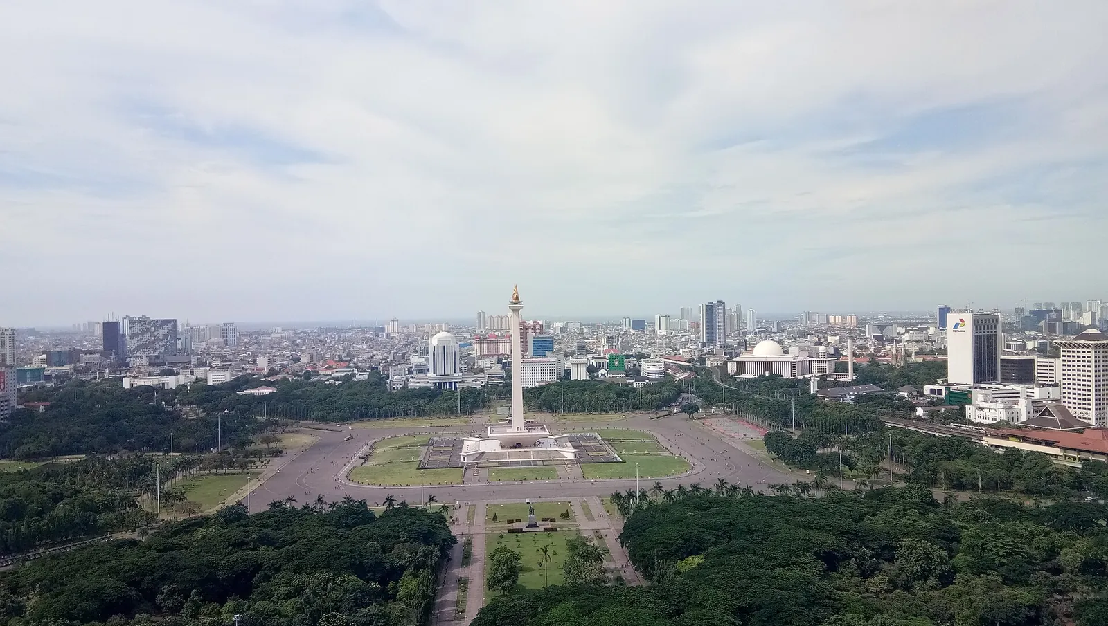 The National Monument (Monas) rising from the green lawns of Merdeka Square in central Jakarta, seen from above with the city spread behind it
