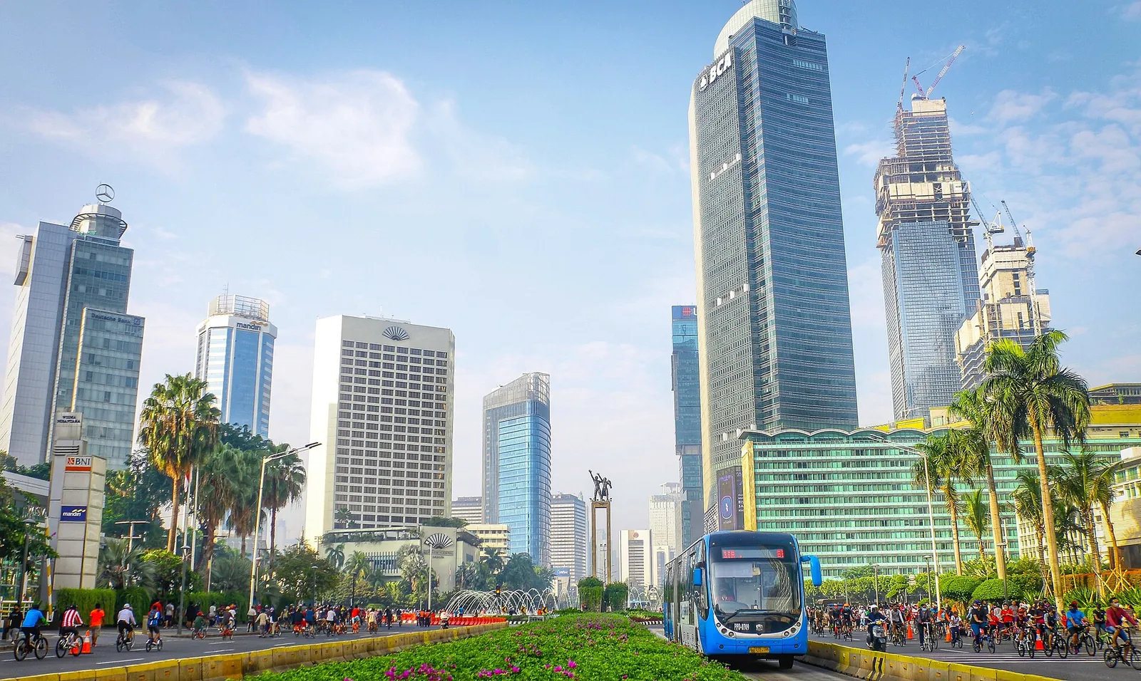 A Transjakarta bus on its dedicated lane at Bundaran HI, with office towers and palm trees under a blue sky