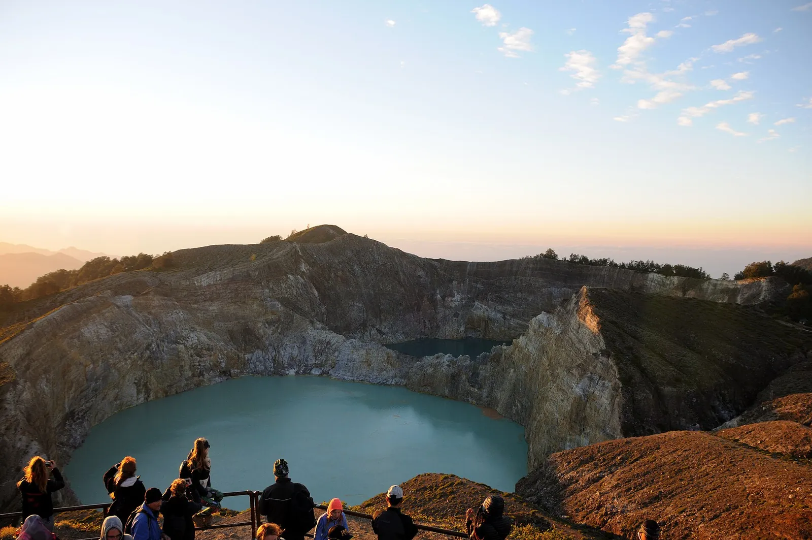 Visitors at a railing looking over Kelimutu's twin crater lakes, a pale turquoise lake in front and a darker one behind, with the sun low on the horizon