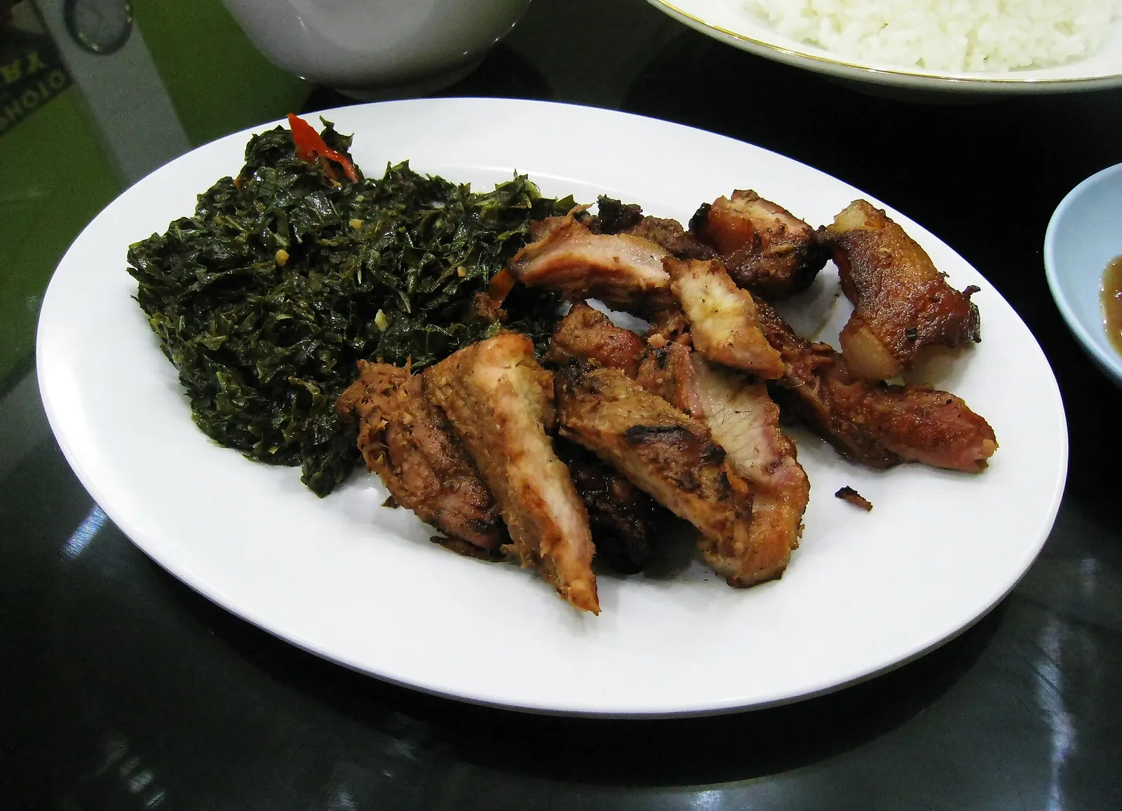A white plate of sliced smoked pork se'i next to sautéed greens, with a bowl of rice and a small dish of sauce at the edges