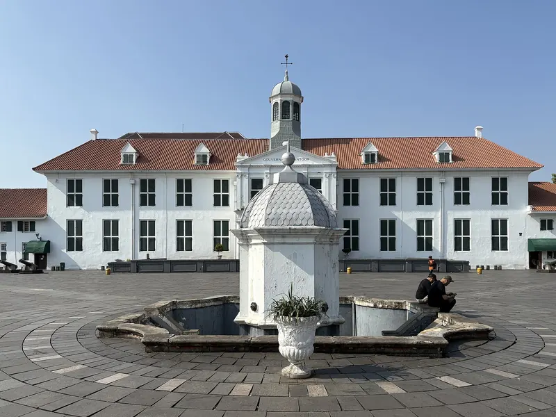 The white former Batavia City Hall, now the Jakarta History Museum, behind the domed fountain on Fatahillah Square under a blue sky