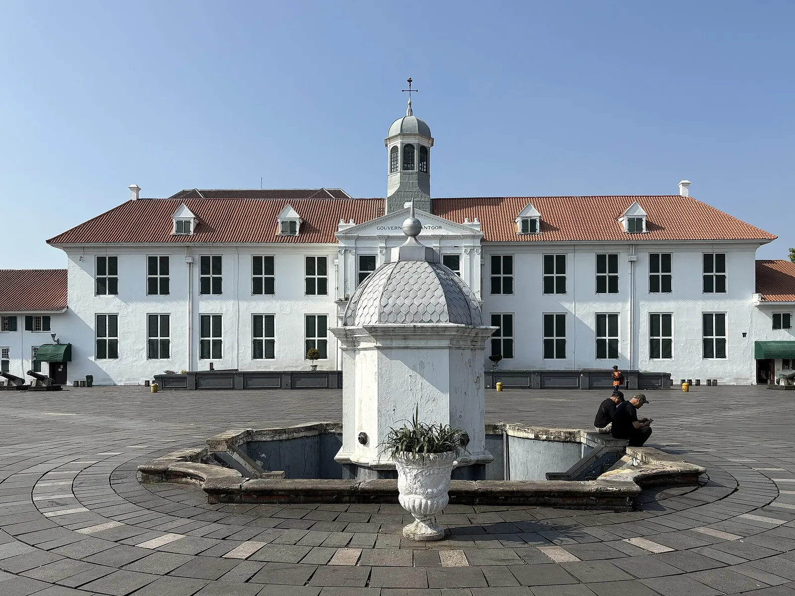 The white former Batavia City Hall, now the Jakarta History Museum, behind the domed fountain on Fatahillah Square under a blue sky