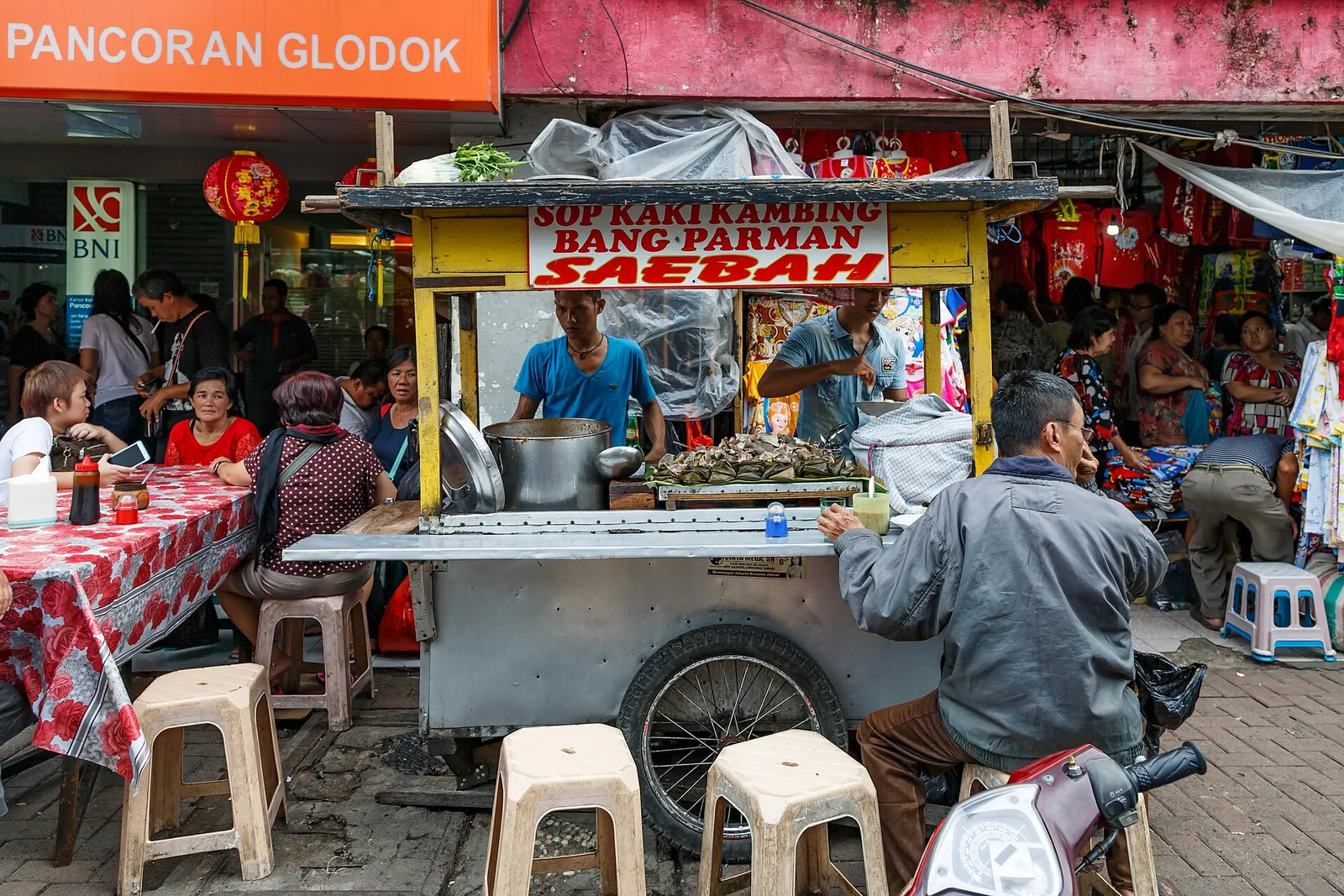 A street stall serving goat feet soup under red lanterns in Glodok, with customers on plastic stools
