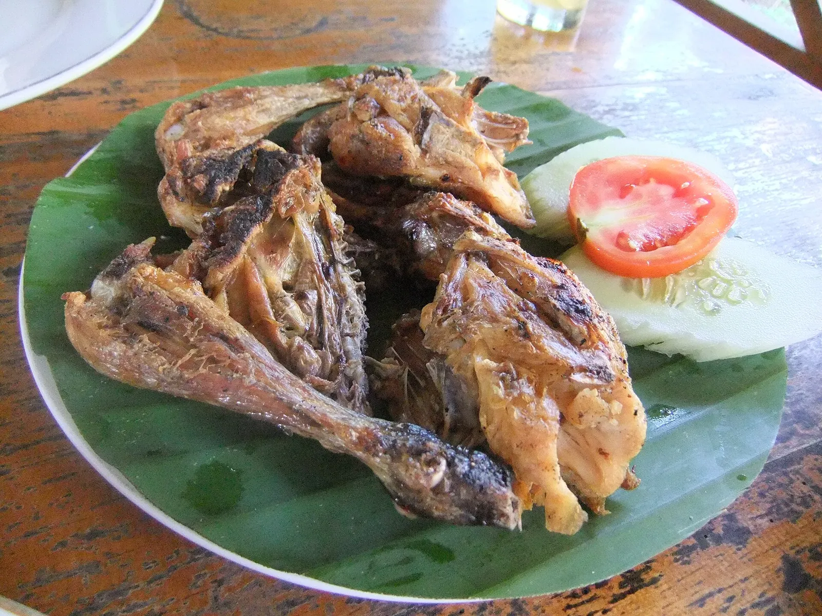 Grilled taliwang chicken on a banana leaf with sliced tomato and cucumber