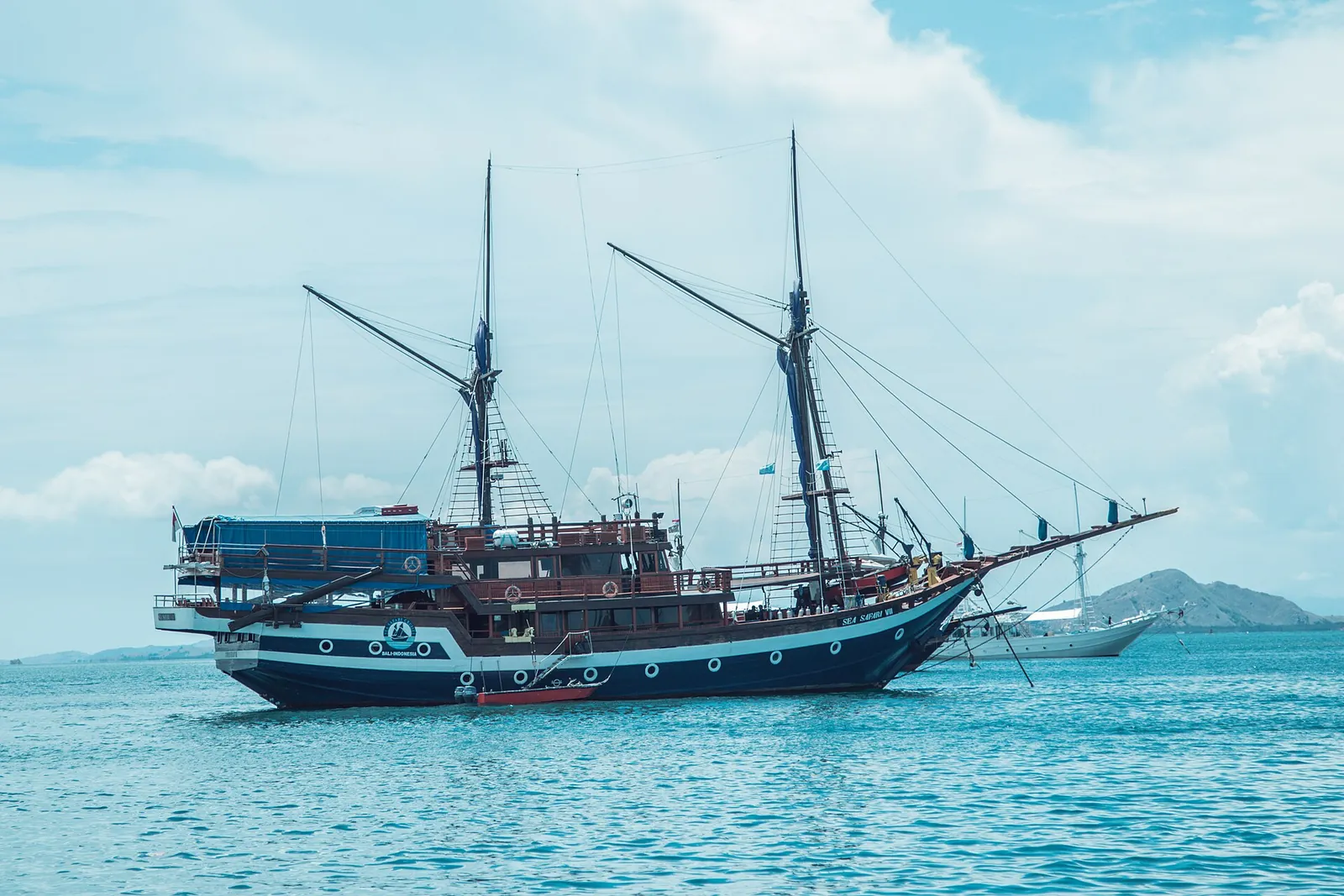 A two-masted wooden phinisi boat at anchor on blue water with a low island behind
