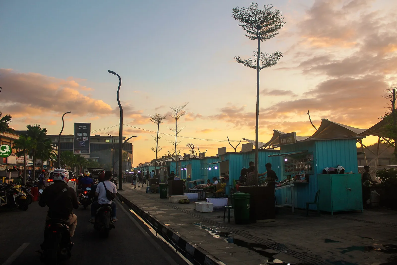 Turquoise food stalls along the seafront road at Kampung Ujung at sunset, with motorbikes passing