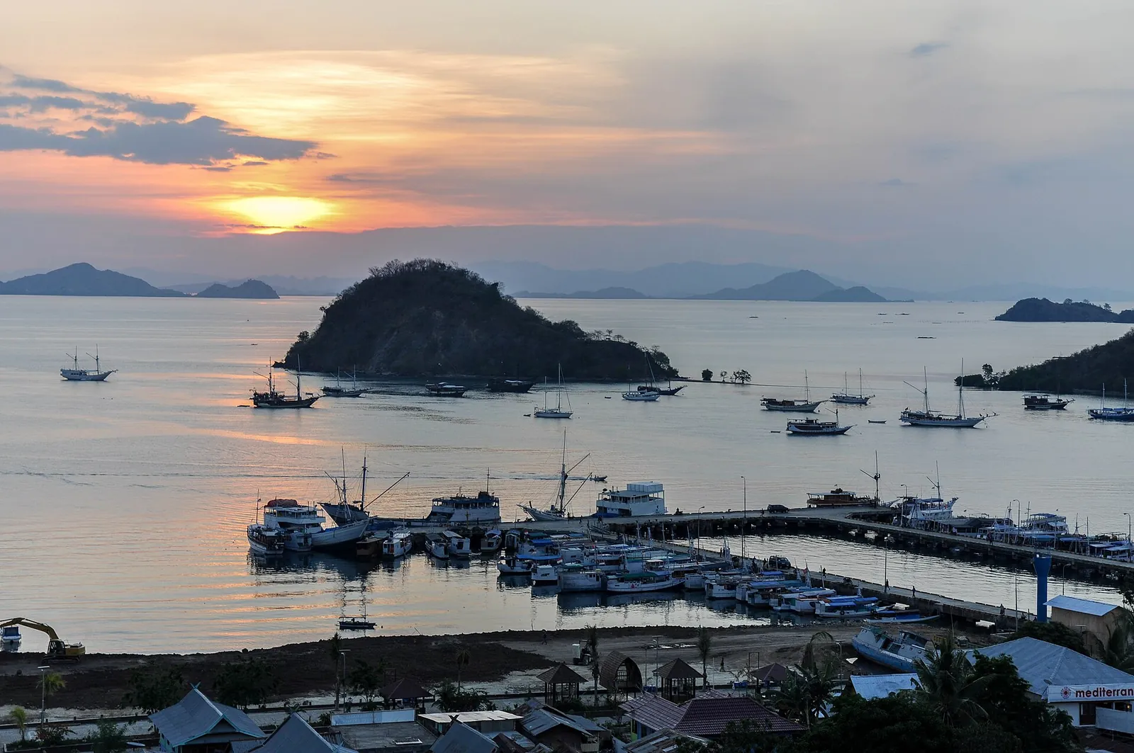 Labuan Bajo's marina and moored boats under an orange sunset sky, with a rounded island beyond