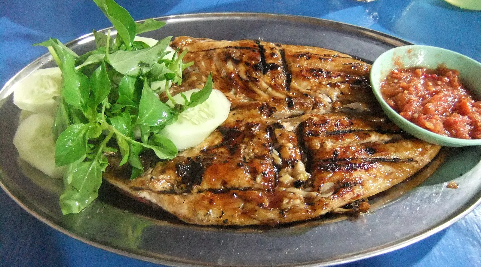 A grilled fish fillet with sambal, cucumber slices and fresh basil on a metal plate