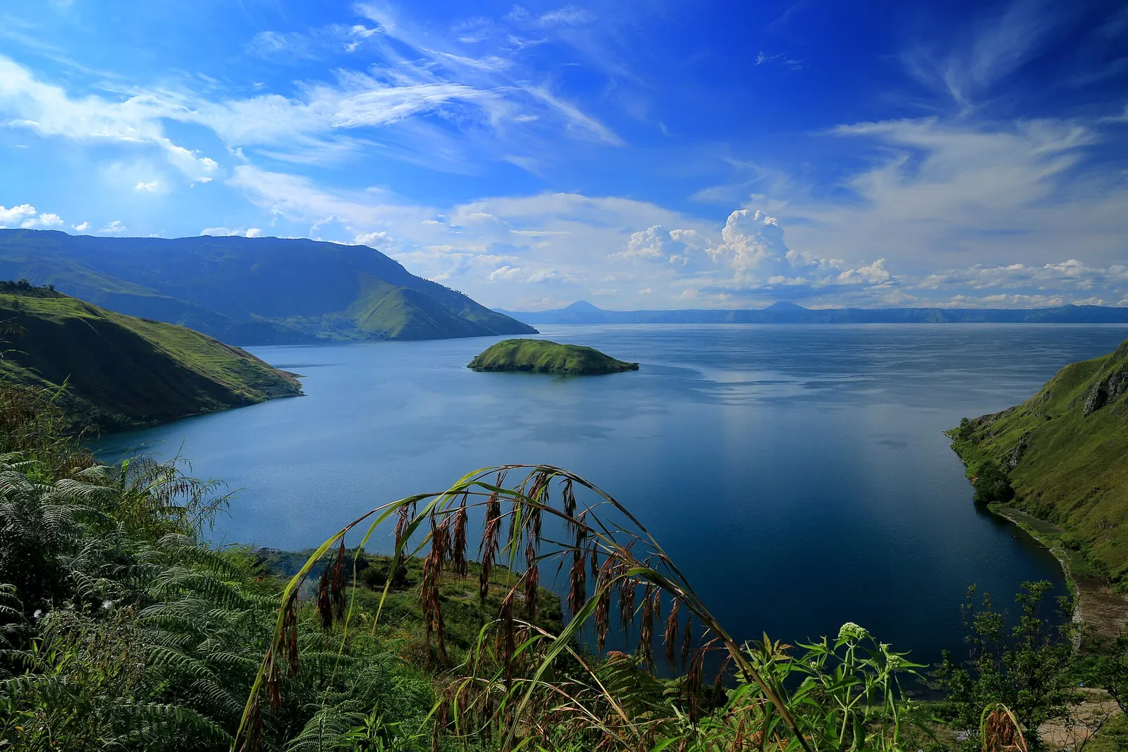 Blue water of Lake Toba between green headlands and a small island, seen from a hillside on Samosir