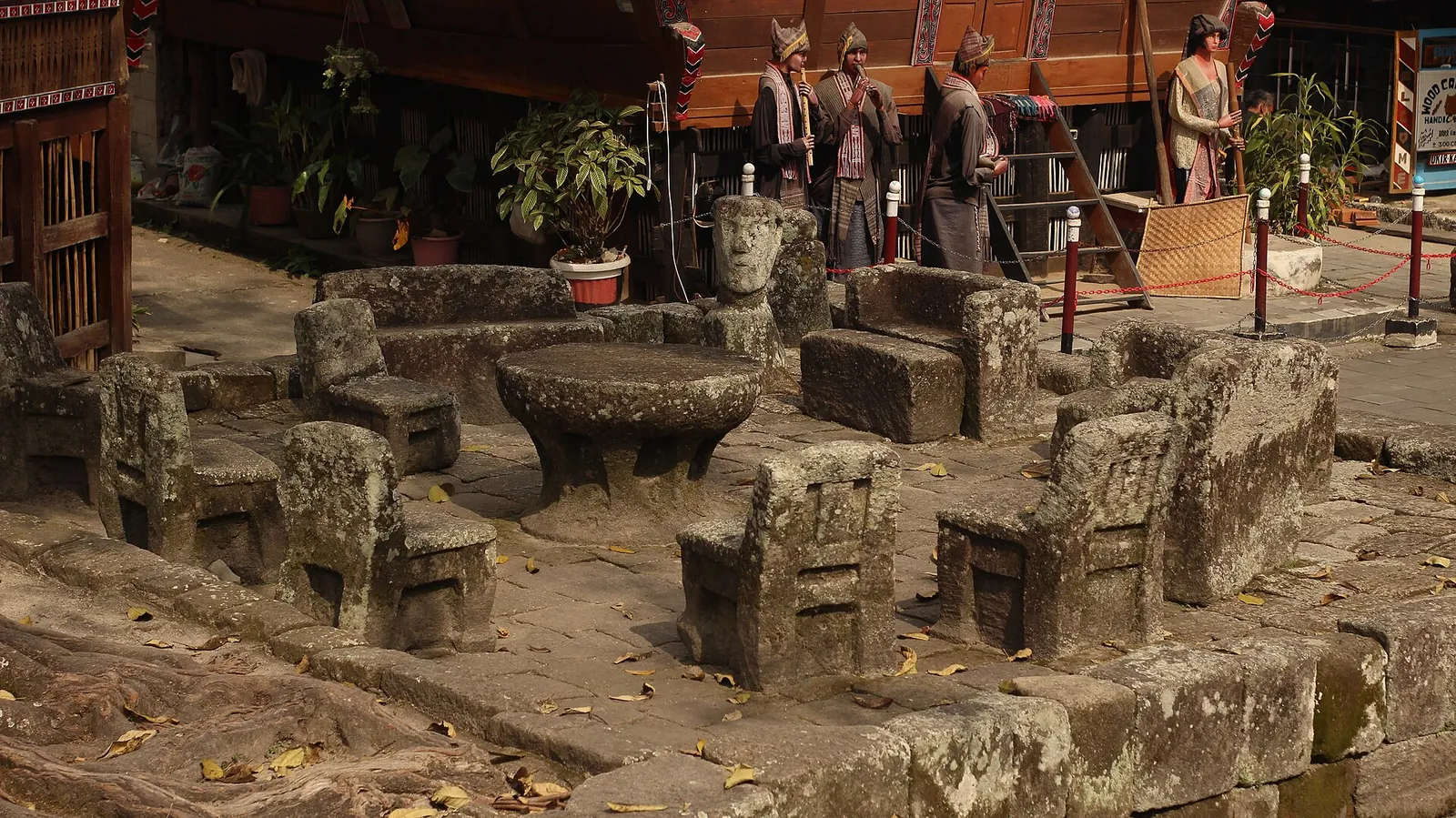 Circle of carved stone chairs around a stone table in the courtyard of Huta Siallagan, with Batak figures behind