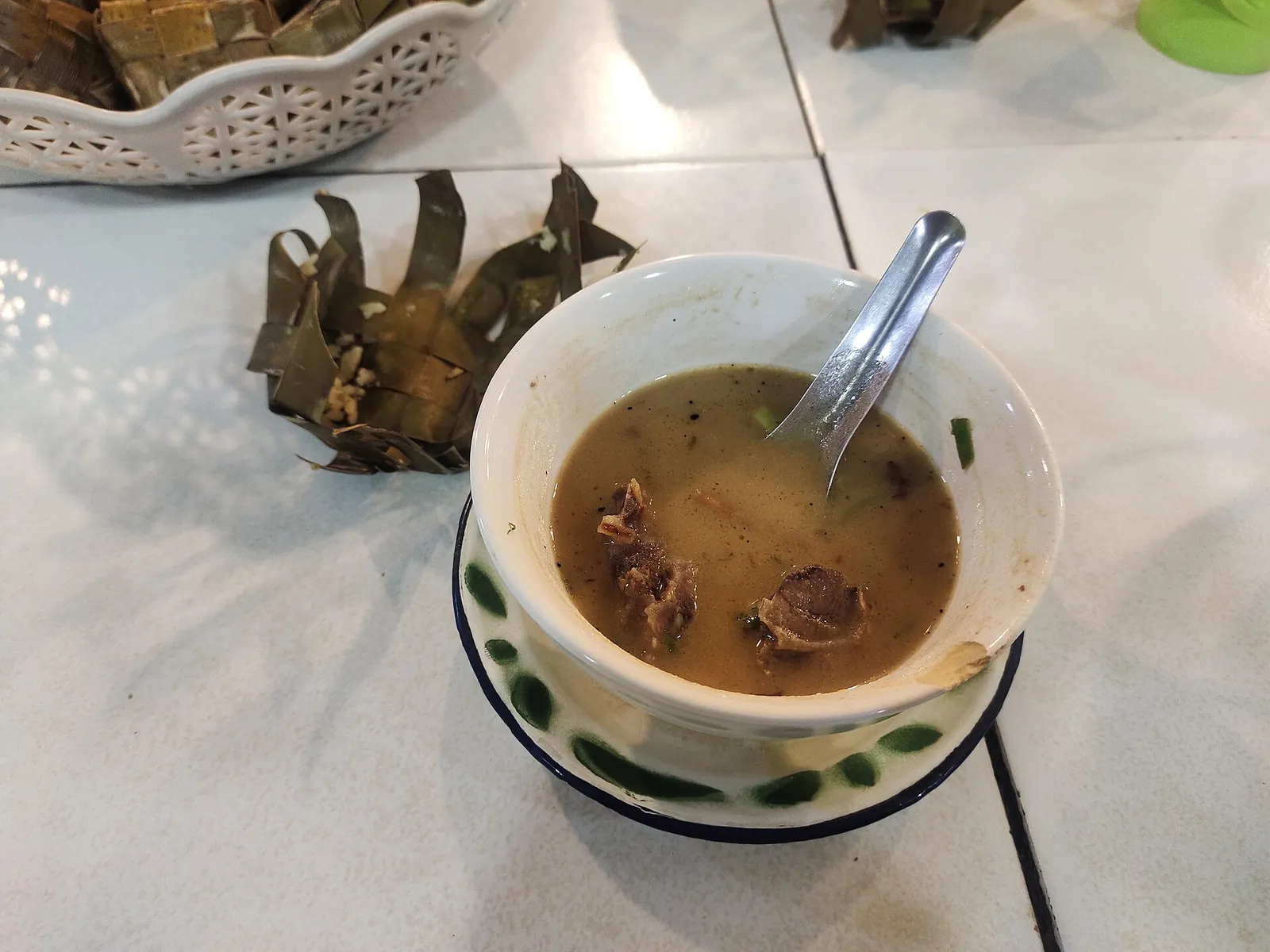 A bowl of coto Makassar, a dark spiced beef soup, with ketupat rice cakes in woven leaves beside it