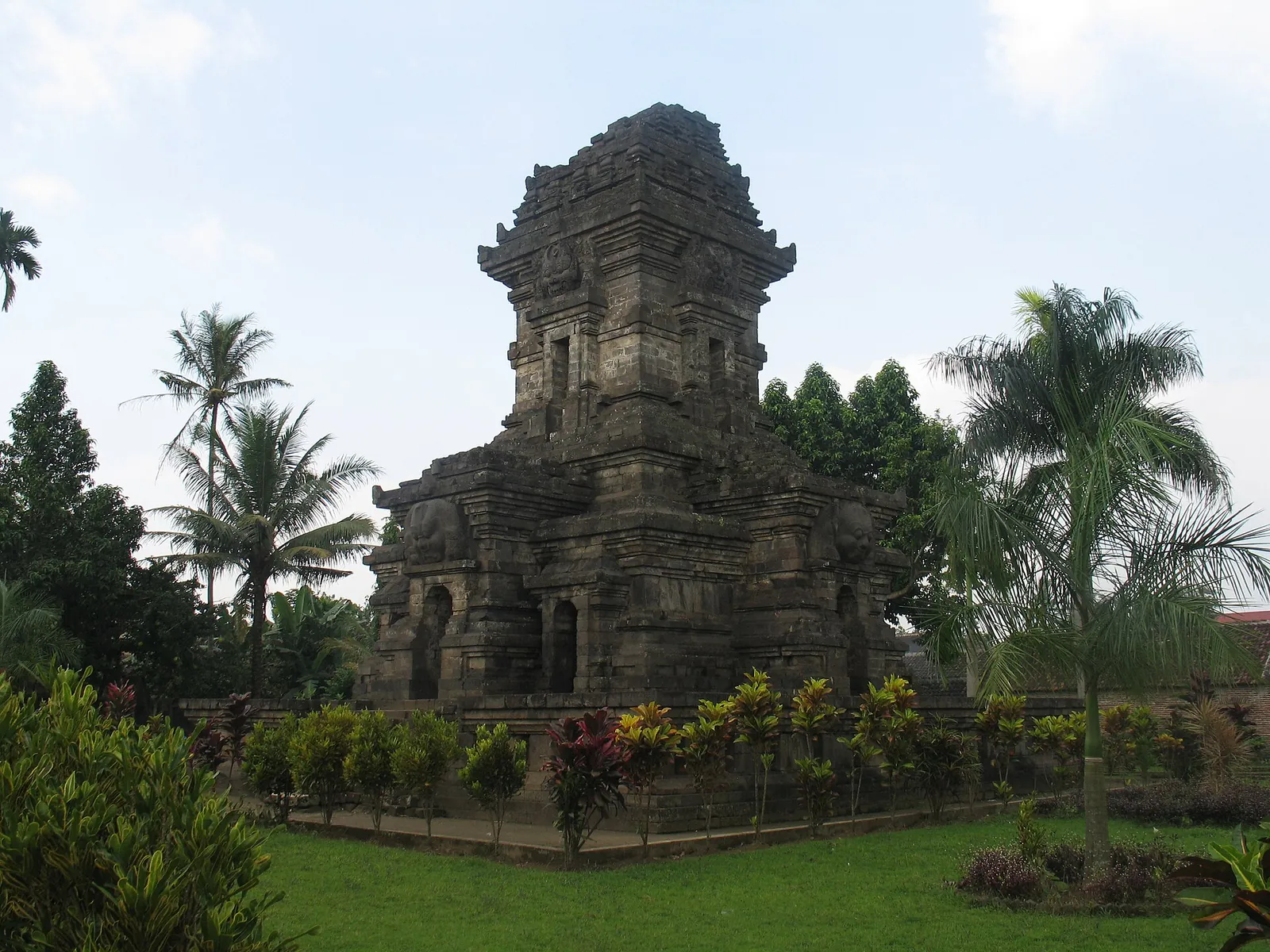 The dark stone tower of Candi Singosari framed by palm trees and a garden