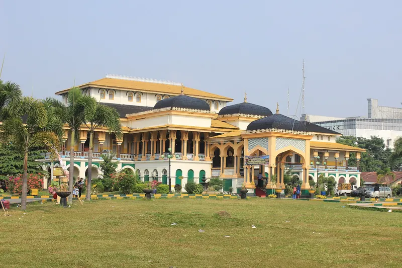 Yellow and green Maimun Palace with dark domed roofs behind a wide lawn and palm trees in Medan