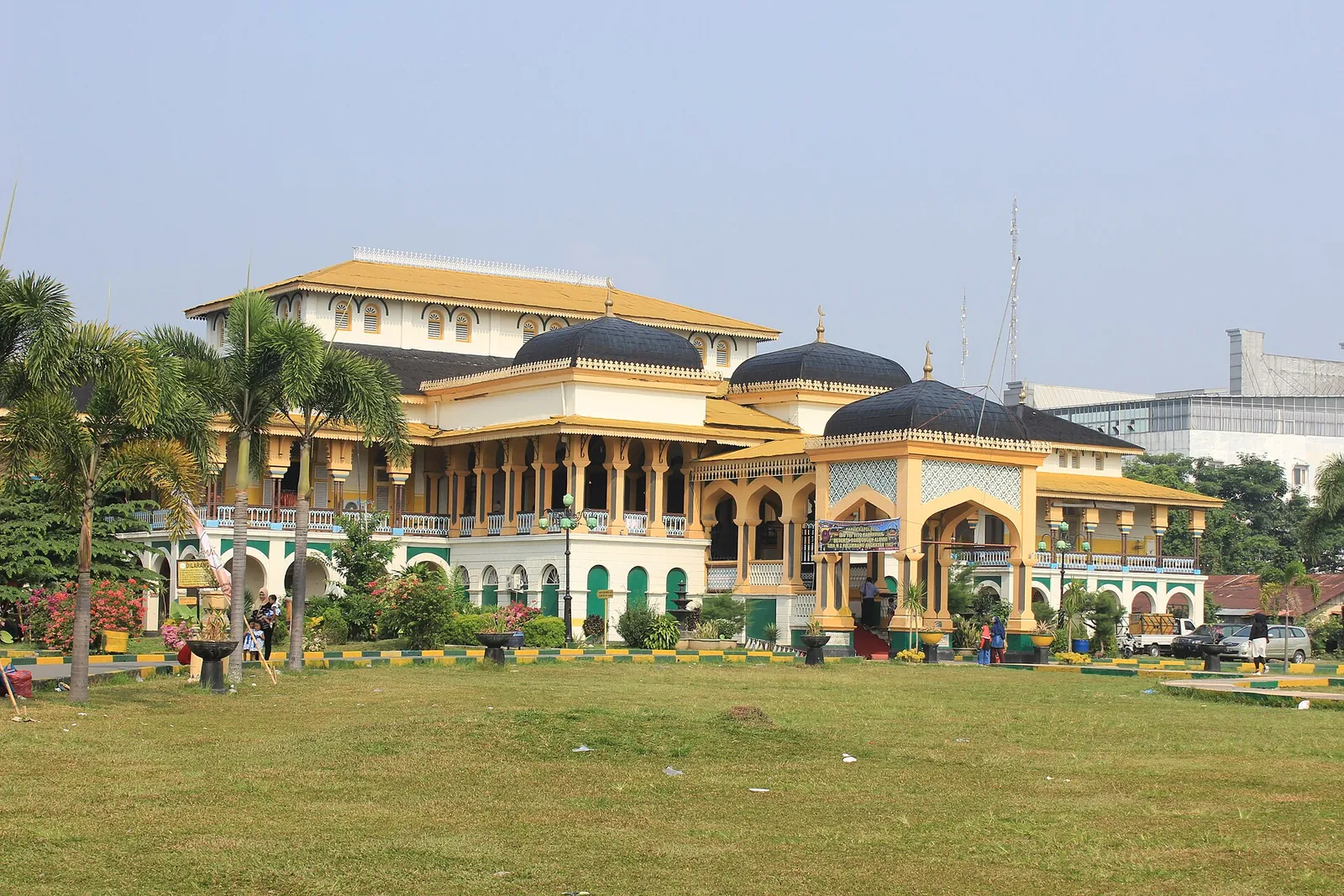 Yellow and green Maimun Palace with dark domed roofs behind a wide lawn and palm trees in Medan