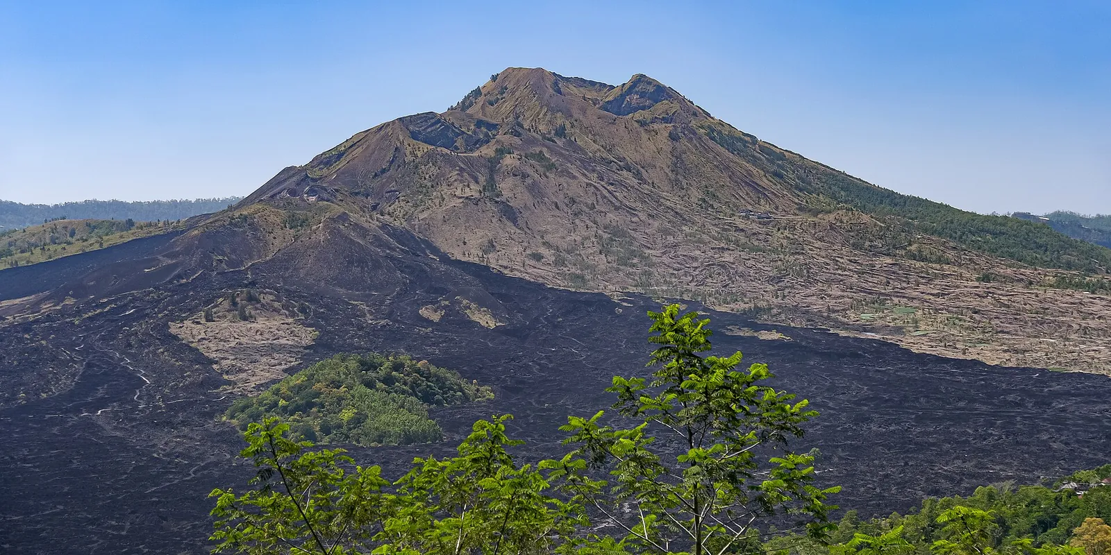 Mount Batur rising above dark lava fields and green forest under a blue sky in Kintamani
