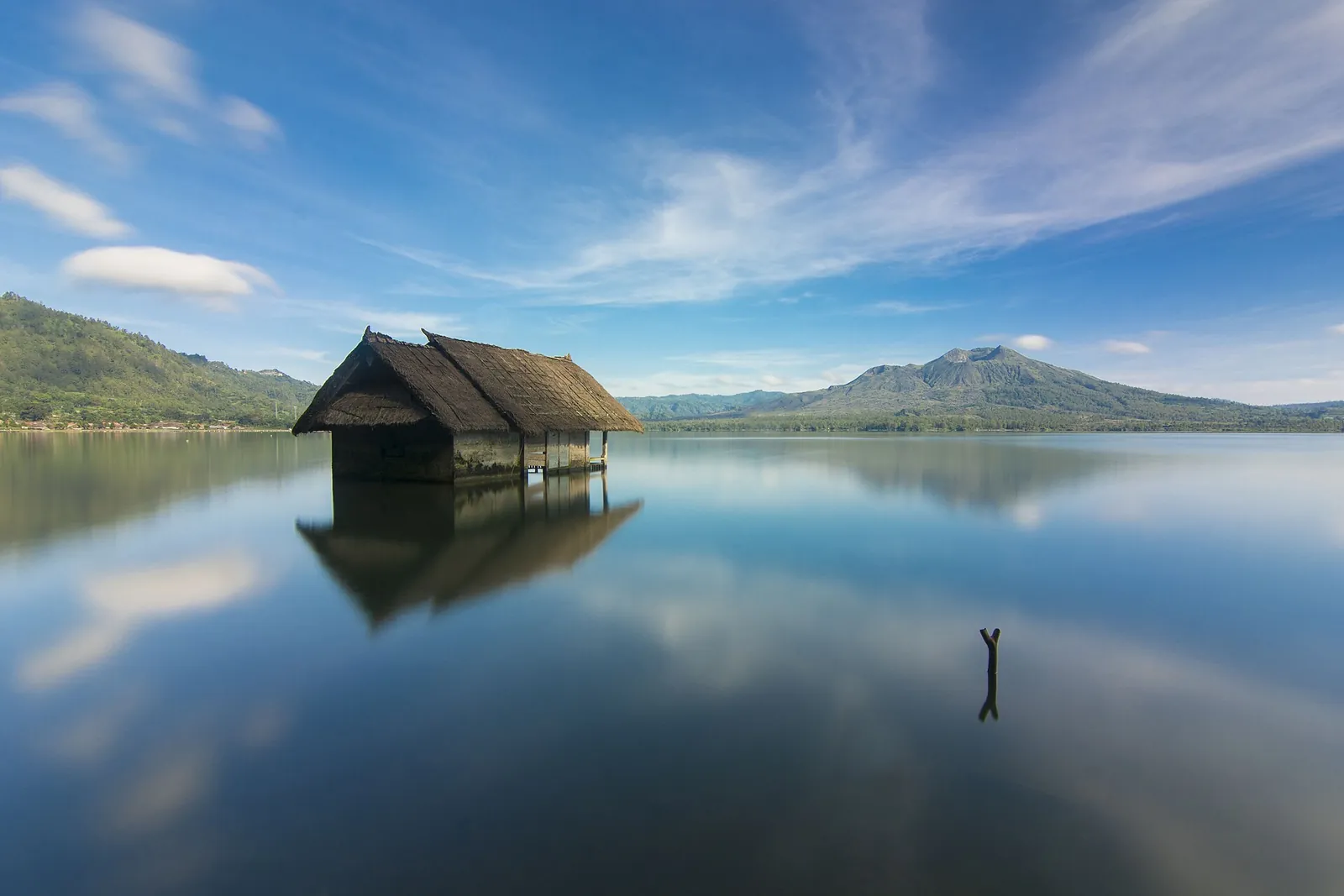 A wooden hut on stilts reflected in the still water of Lake Batur, with hills behind