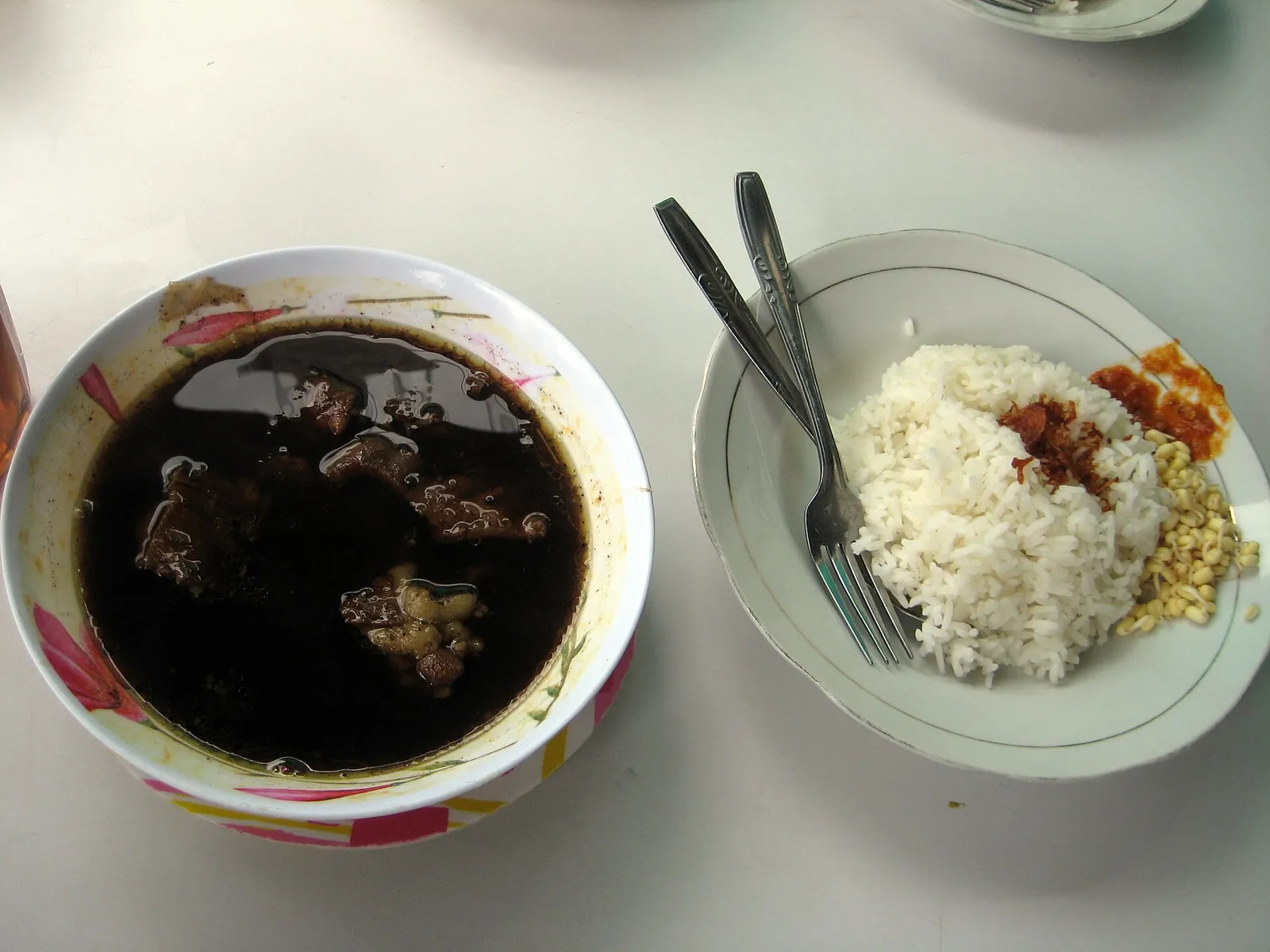A bowl of dark rawon beef soup beside a plate of rice, bean sprouts and sambal at a warung table