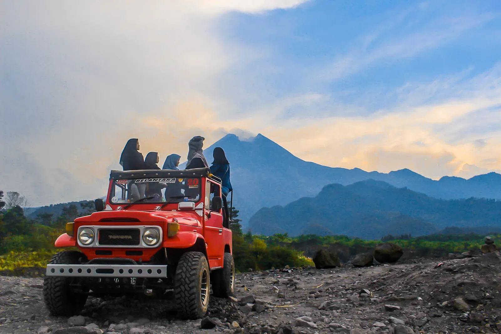 A red open-top jeep carrying tourists on a rocky track with volcanic mountains behind
