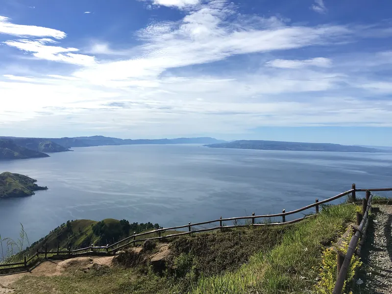 Lake Toba and the shore of Samosir Island seen from a hillside viewpoint with a wooden railing under a blue sky