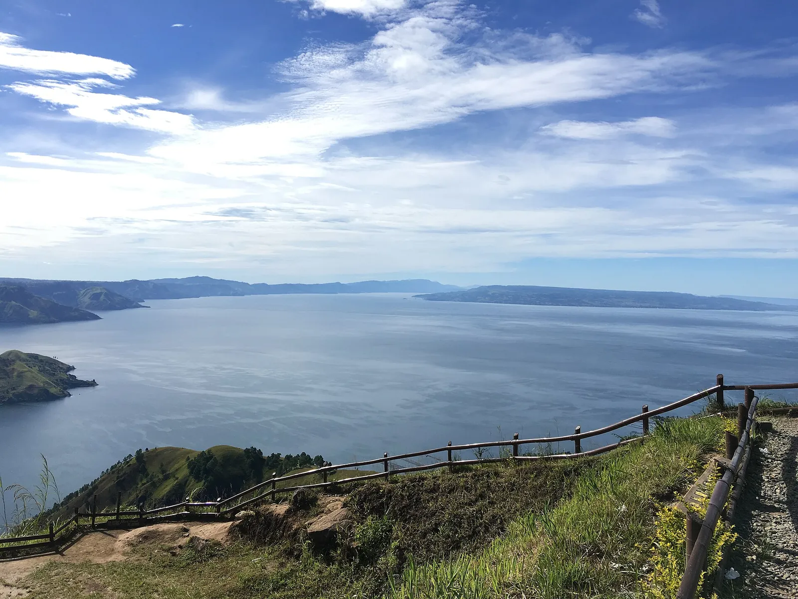 Lake Toba and the shore of Samosir Island seen from a hillside viewpoint with a wooden railing under a blue sky