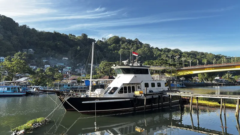 Boats moored on the Batang Arau river in Padang with the yellow arch of the Siti Nurbaya Bridge and wooded hills behind