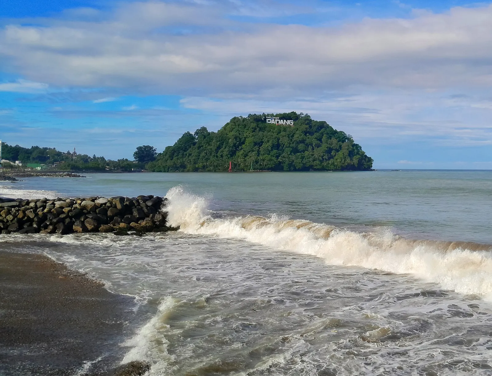 Waves breaking on a breakwater at Padang Beach with the wooded hill of Gunung Padang and its PADANG sign offshore
