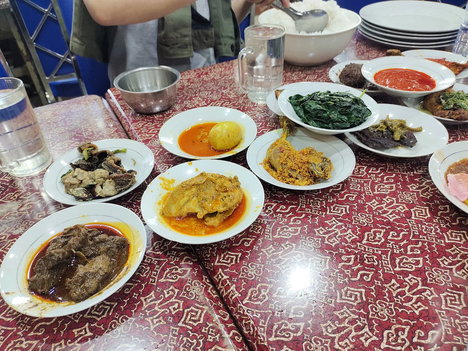 A table of small plates of nasi padang dishes, including rendang, curries, greens and sambal, around a bowl of rice