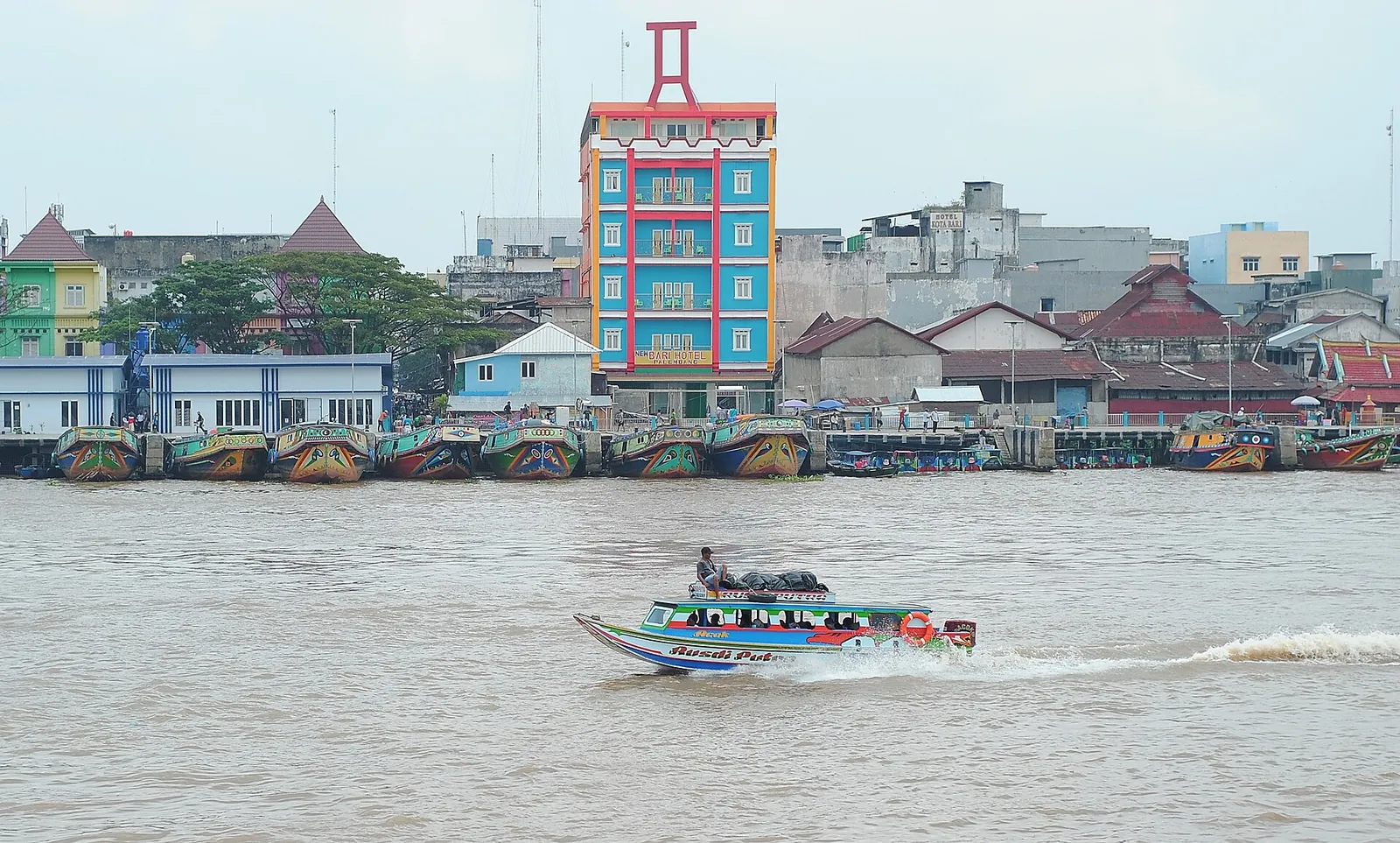 A speedboat crossing the Musi River in front of colourful riverside buildings and moored boats in Palembang