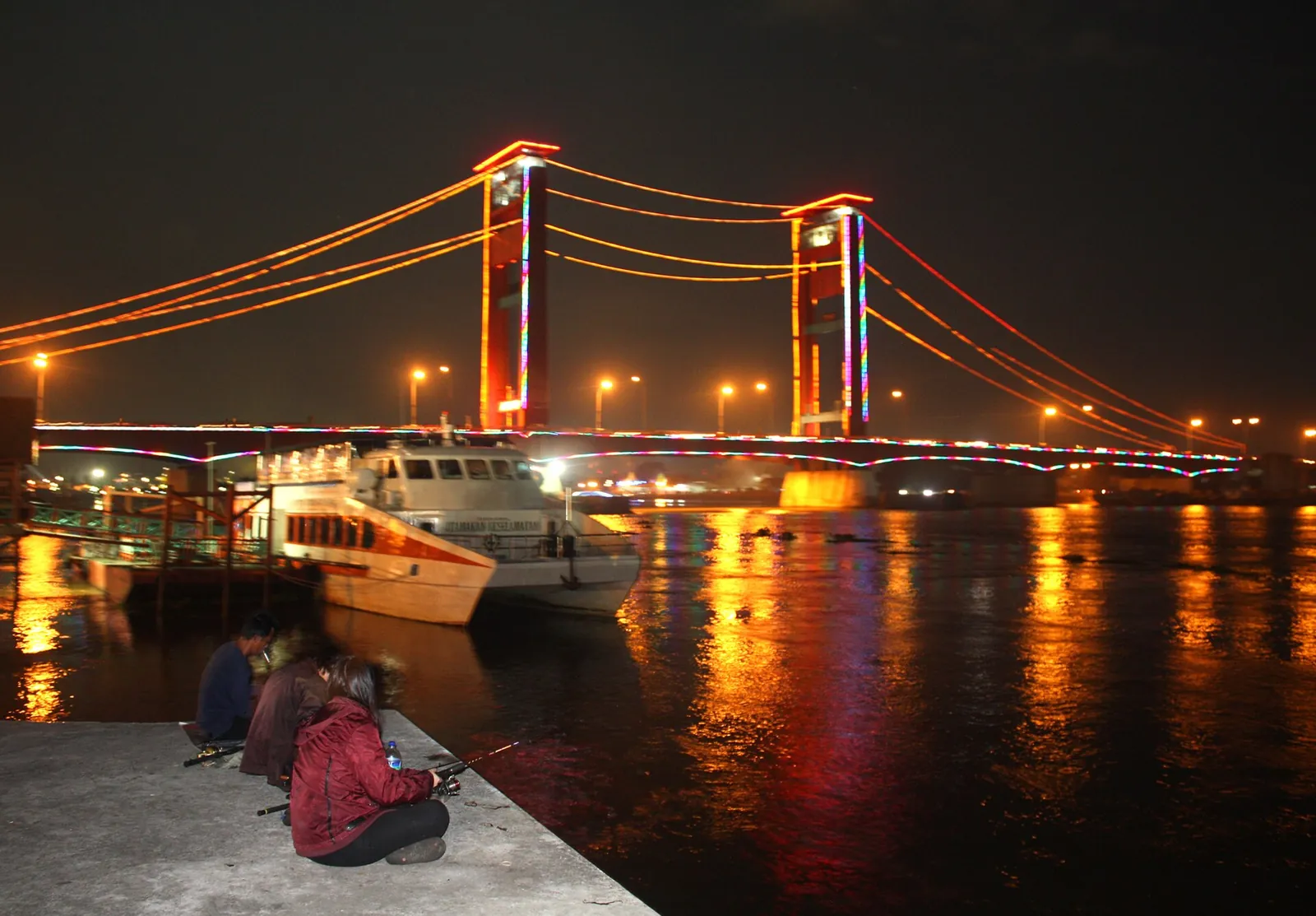 The Ampera Bridge lit up in red and orange at night, with lights reflected in the Musi and people sitting on the riverbank