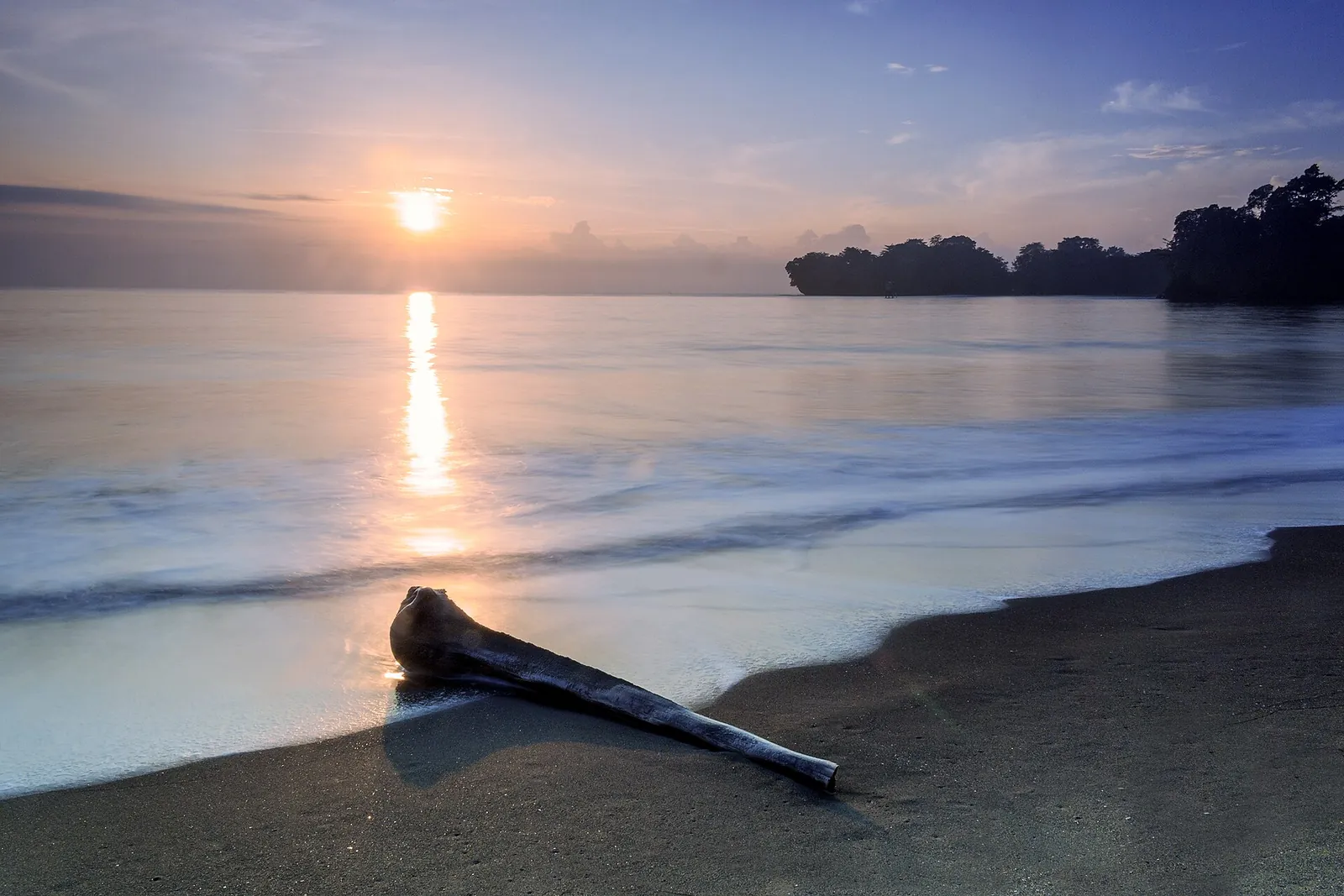 The sun rising over a calm sea and dark sand at Batu Karas, with a rocky wooded point on the horizon