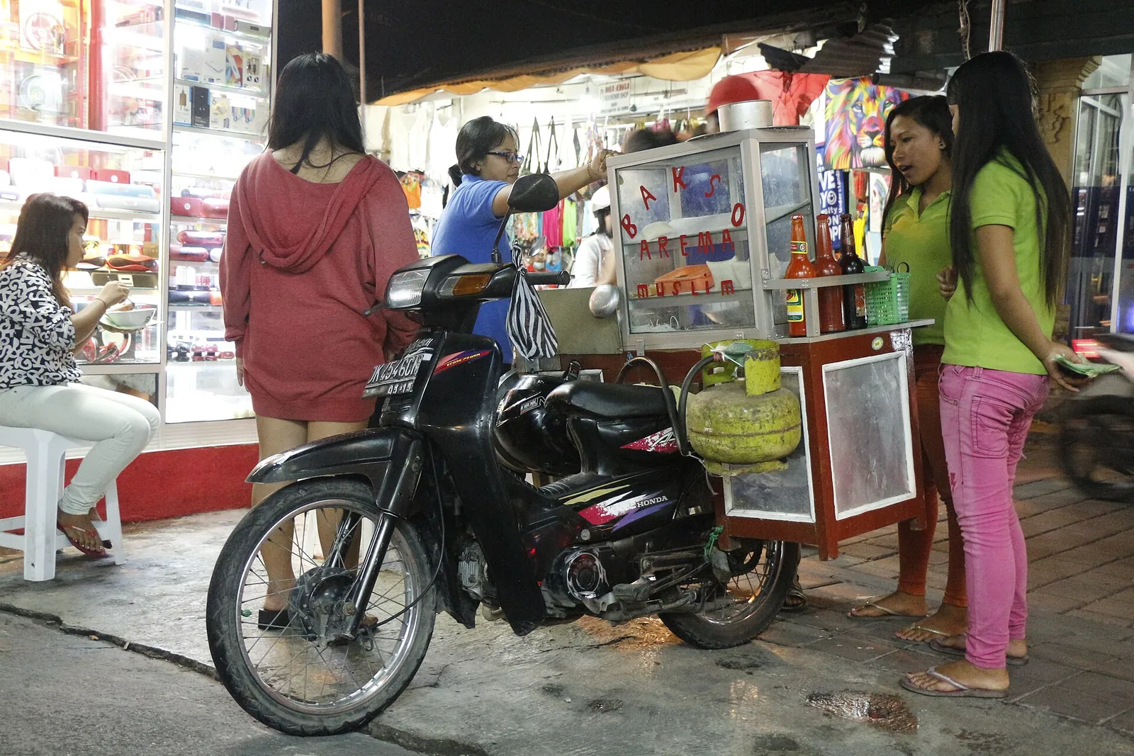 A motorbike bakso cart parked on a pavement in Kuta at night, with customers waiting beside it