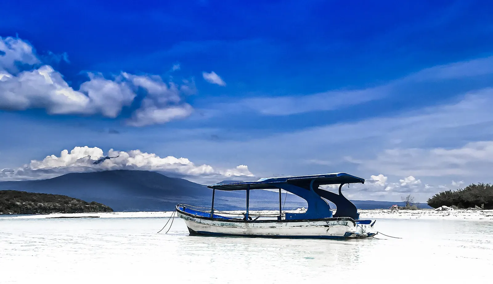 A covered wooden boat moored in clear turquoise shallows off Menjangan Island, with mountains across the water