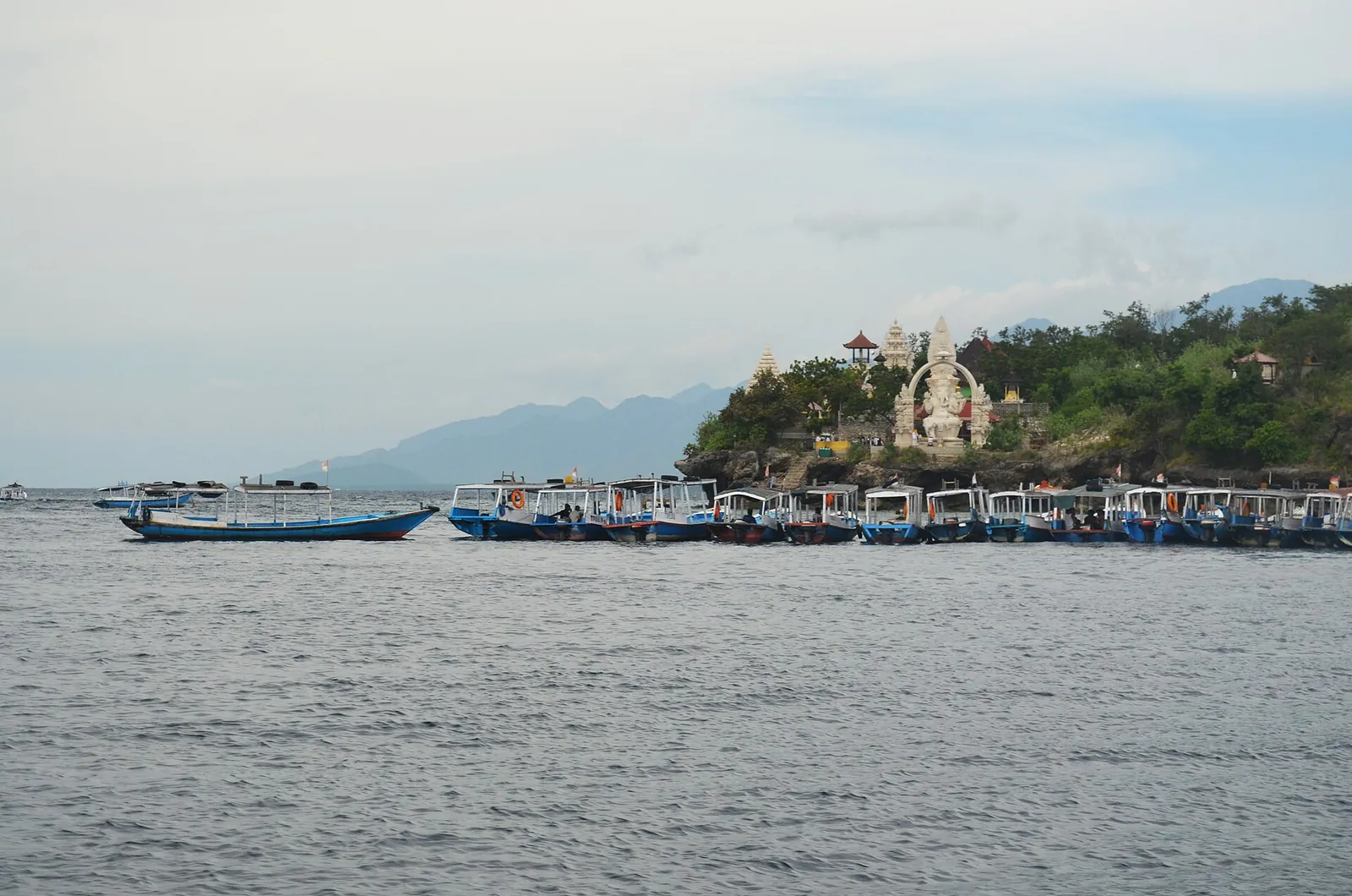 Tourist boats lined up off Menjangan Island below a hilltop temple with a large white statue