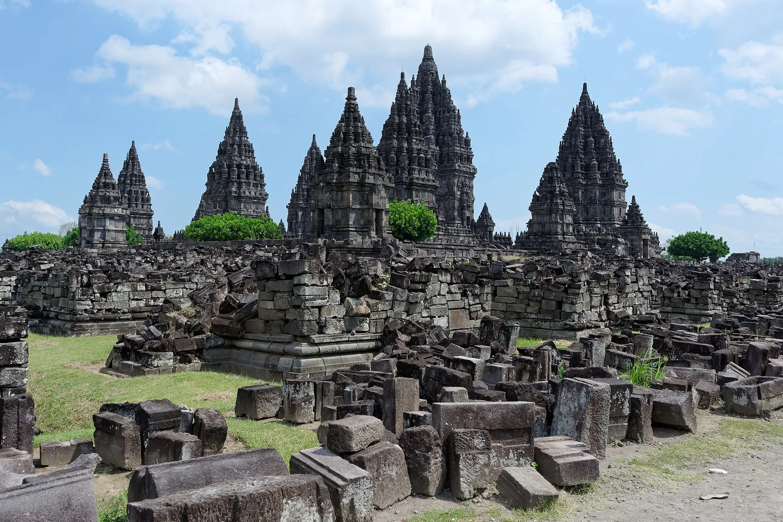 The tall shrines of Prambanan rising above fields of fallen stone blocks under a blue sky