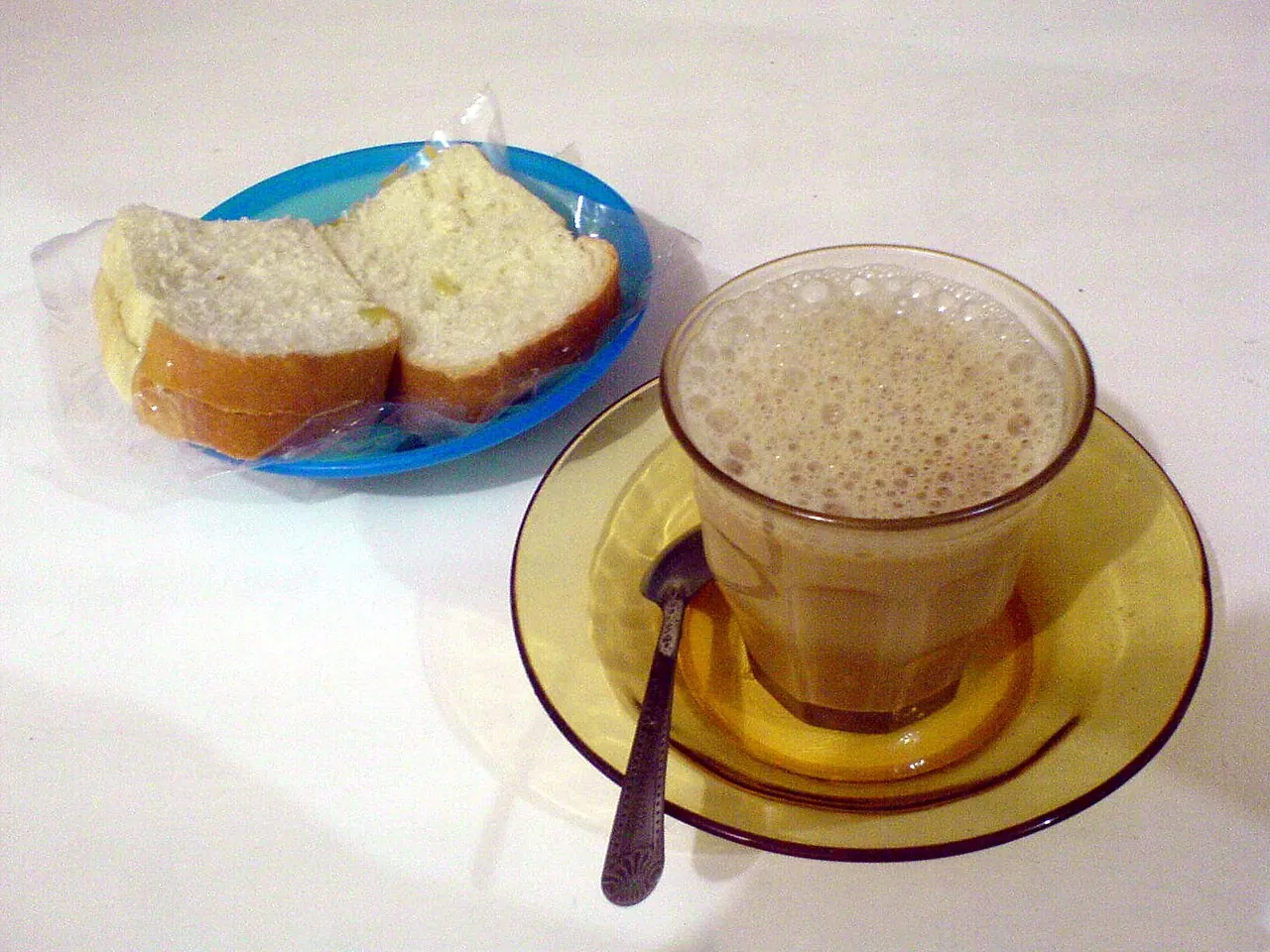 A glass of frothy kopi sanger on a saucer next to slices of bread with spread